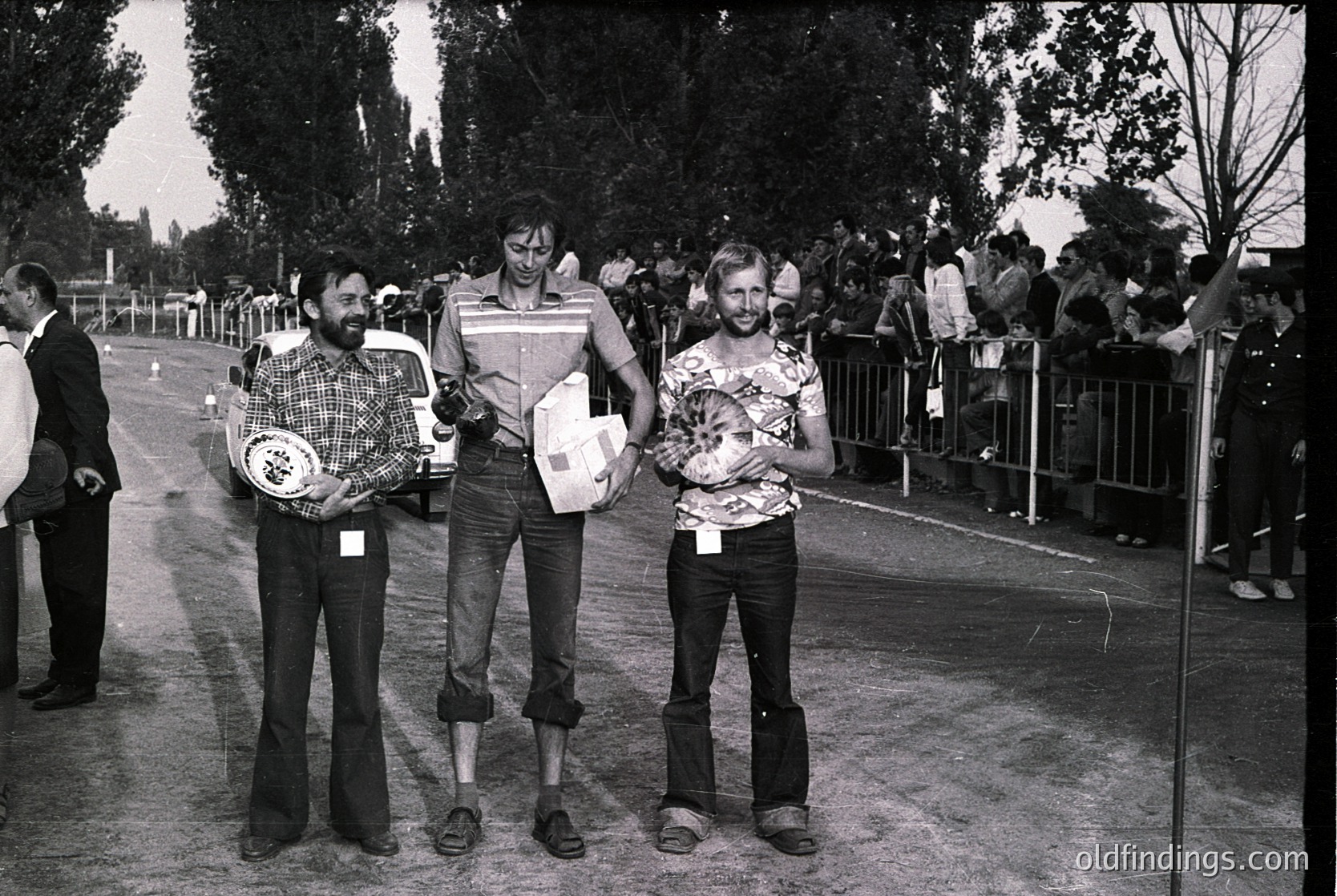 Three men pose holding framed plaques at an outdoor event, likely a sports or cultural competition. The central figure wears a checkered shirt and holds a rectangular plaque with text. Surrounding spectators stand behind barriers. Mid-20th century attire and setting suggest or . Crowd and barriers indicate organized event atmosphere.