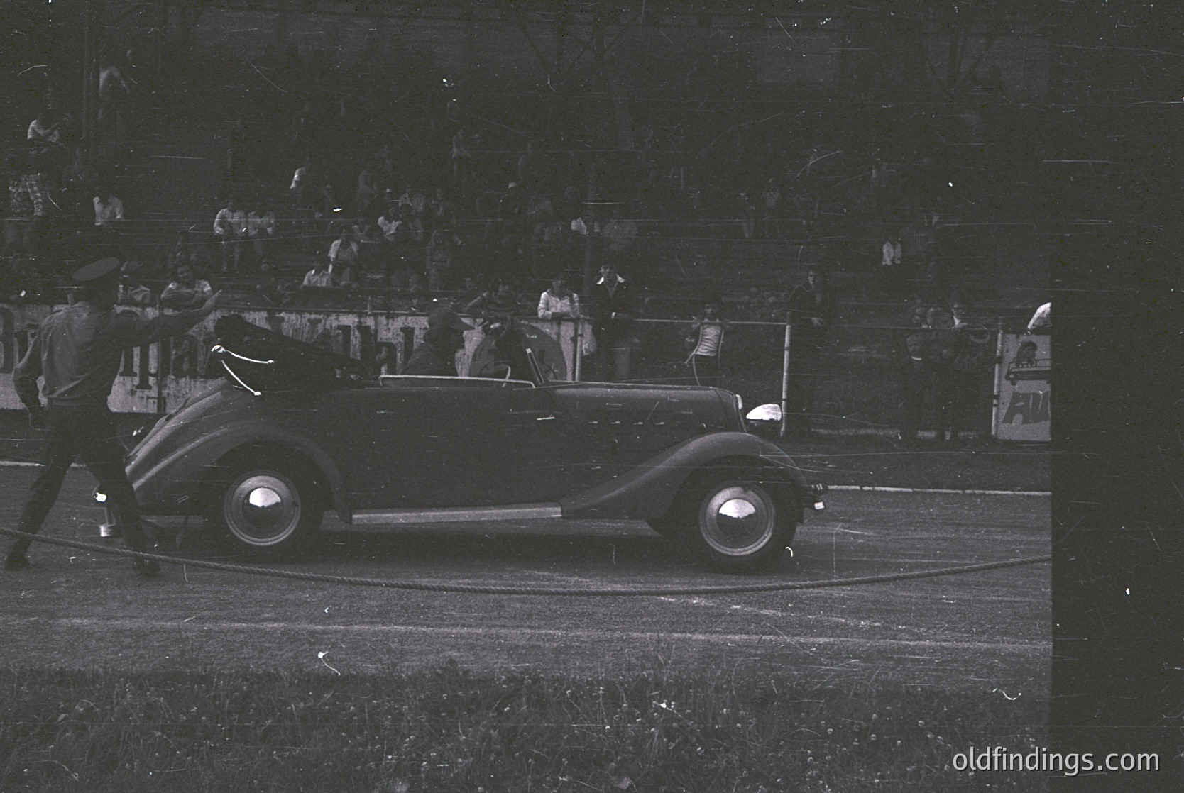 Classic 1930s convertible race car on a track, surrounded by spectators. Streamlined bodywork and dual headlights highlight vintage automotive design. Crowd in stands suggests motorsport event.