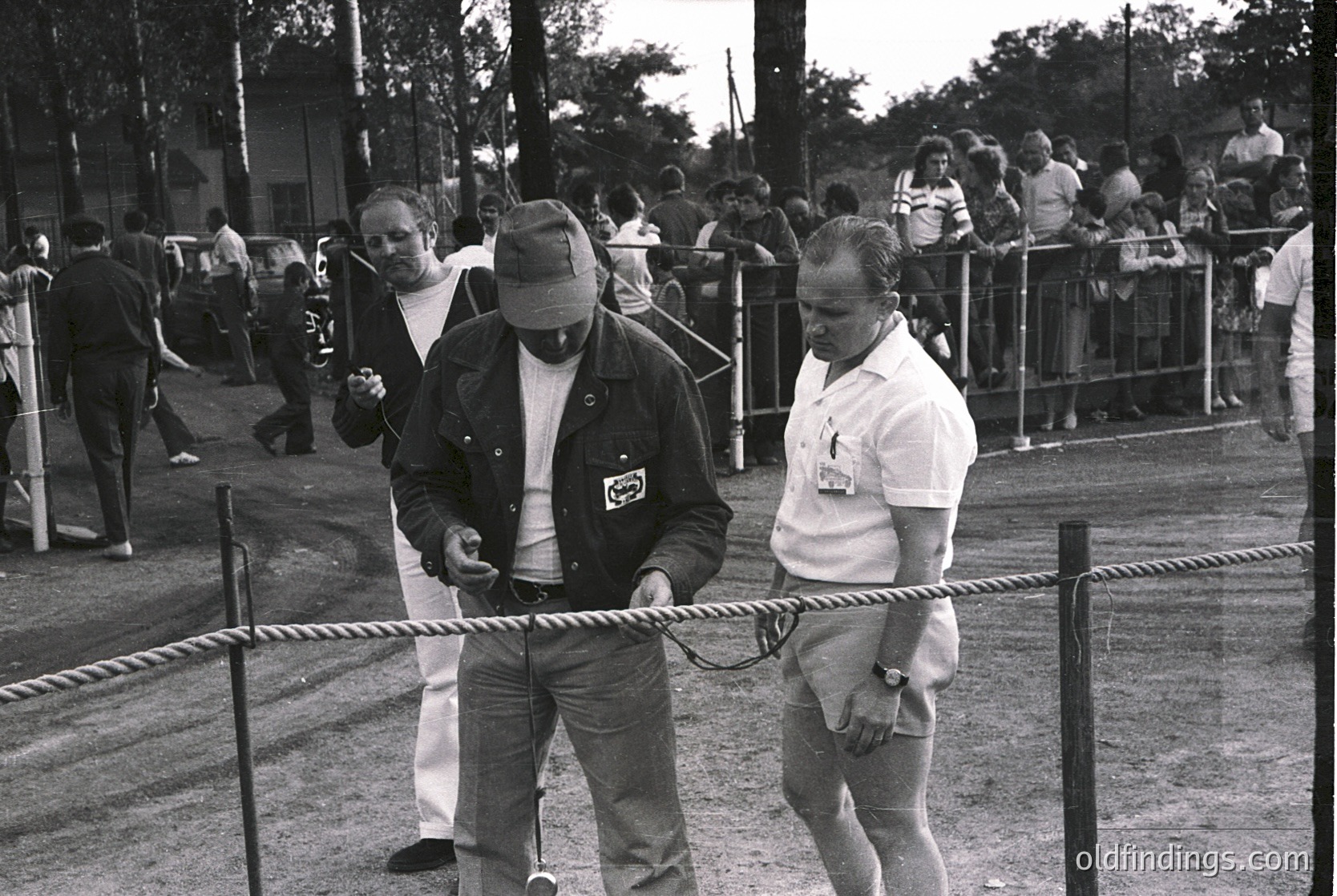 Mid-20th century outdoor event with rope barrier separating crowd from participants. Two men in casual workwear (one in a hat, leather jacket, and lanyard; the other in a white shirt and shorts) guide a rope barrier. Spectators in casual attire stand behind metal fencing. Likely a public gathering or festival. -1970s