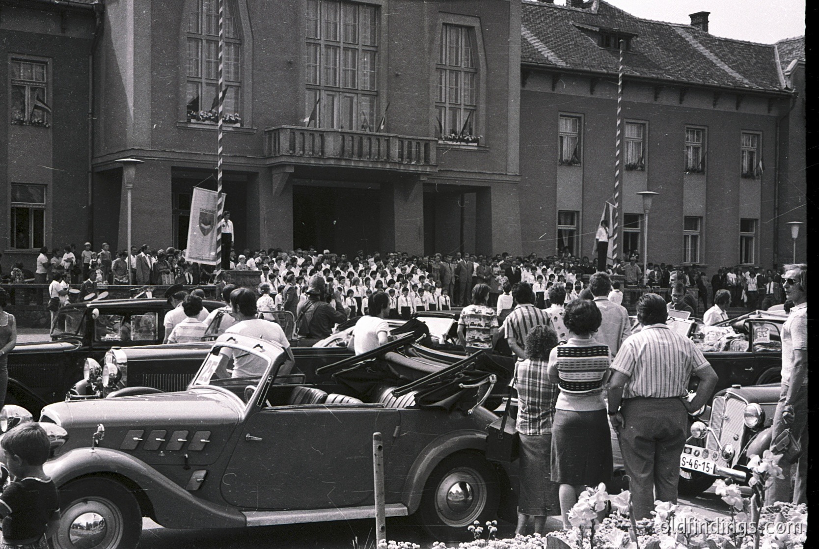 Black-and-white street scene featuring a large crowd gathered around a grand, neoclassical building with arched windows and a balcony. Mid-20th century convertible cars parked in front, with spectators standing and leaning on vehicles. Prominent flags and banners suggest a public event or ceremony. Crowd density indicates a significant gathering, likely in a European city.