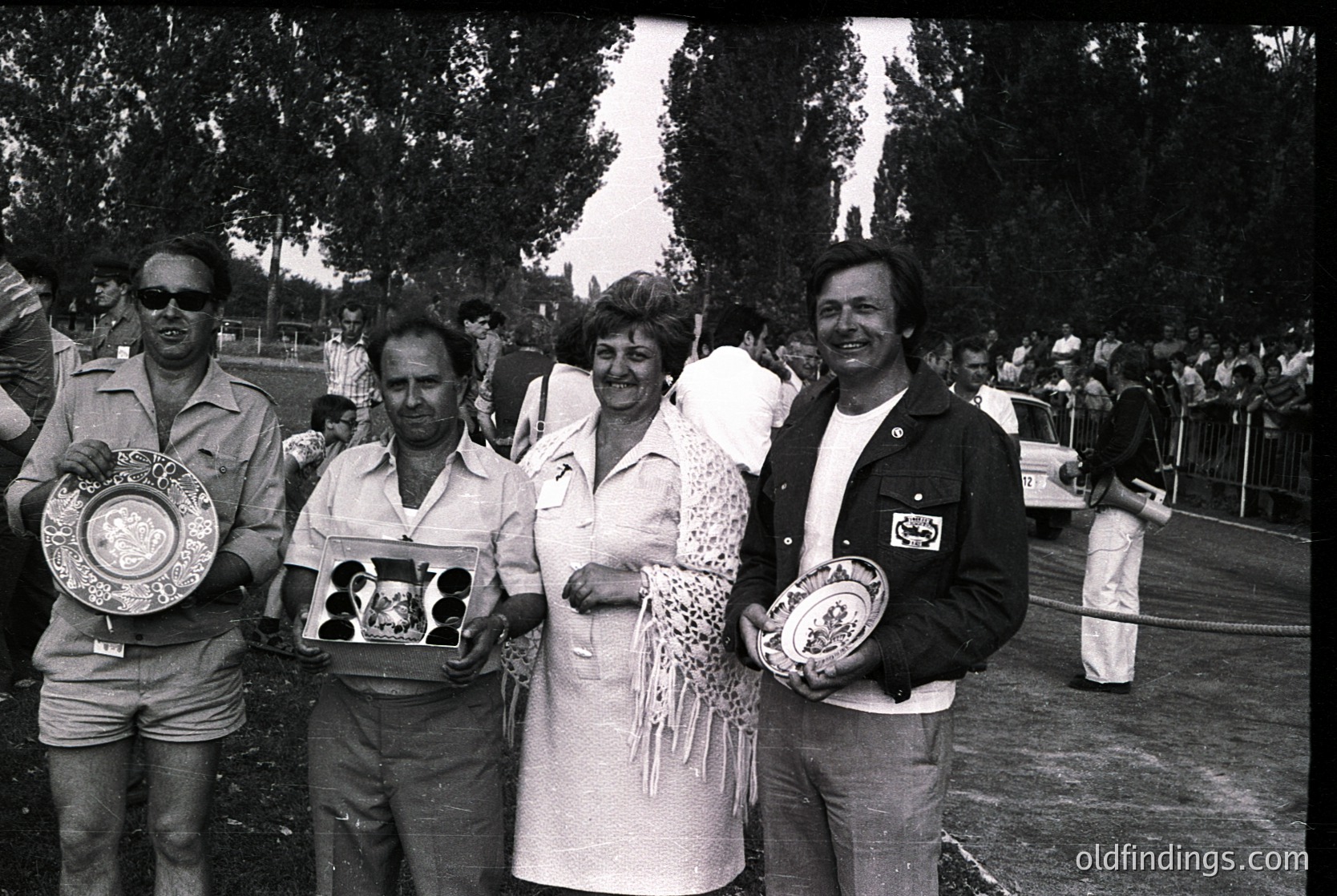 Four individuals pose outdoors holding trophies, likely from a 1970s sports or cultural event. The man on the left wears sunglasses and a khaki jacket; the woman in the center has a patterned shawl. Crowds and barriers suggest a public venue.