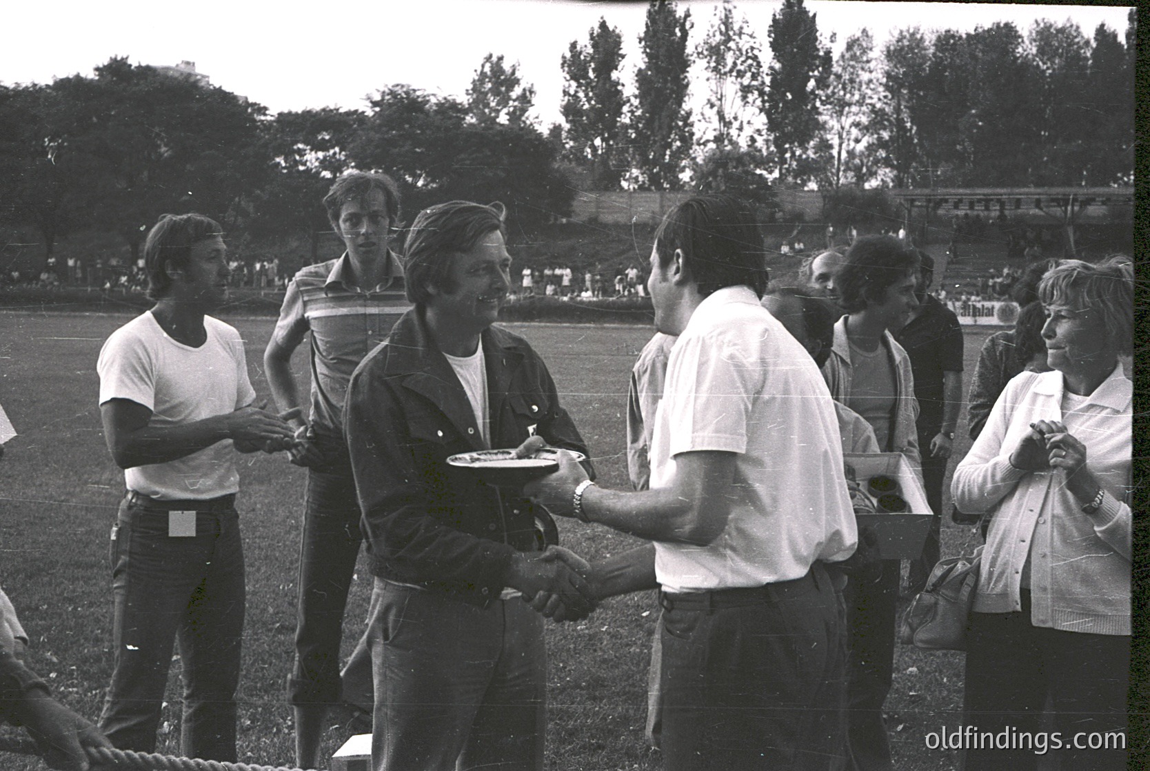 Handshake ceremony on grassy field with blurred stadium in background. Mid-20th century sports event, likely 1960s–1970s. Winner in cap and vest receives medal from official in casual attire.