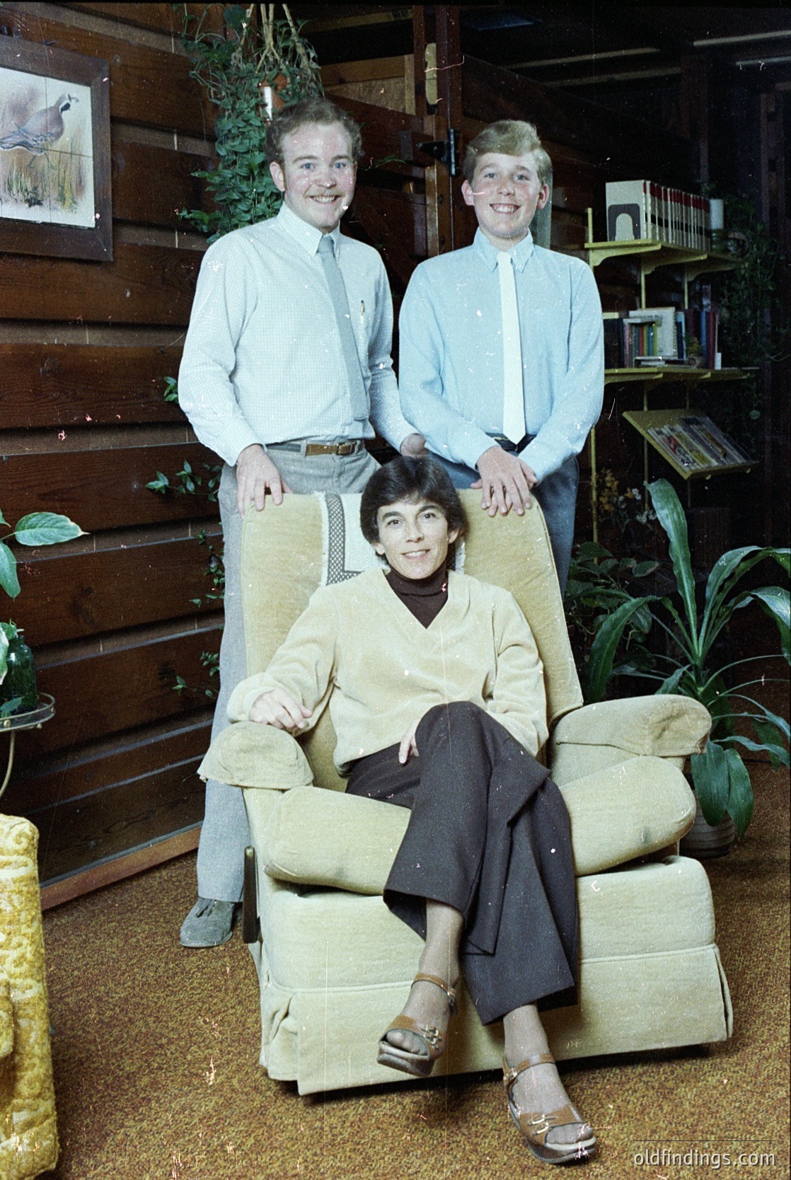 Family portrait in a rustic, wood-paneled interior, likely mid-20th century (1960s–1970s). Three individuals pose: two standing boys in light blue button-down shirts and khaki pants, one seated woman in a beige sweater and dark trousers with sandals. Background features wooden shelves with framed art, potted plants, and a bird illustration. Warm, homely atmosphere.