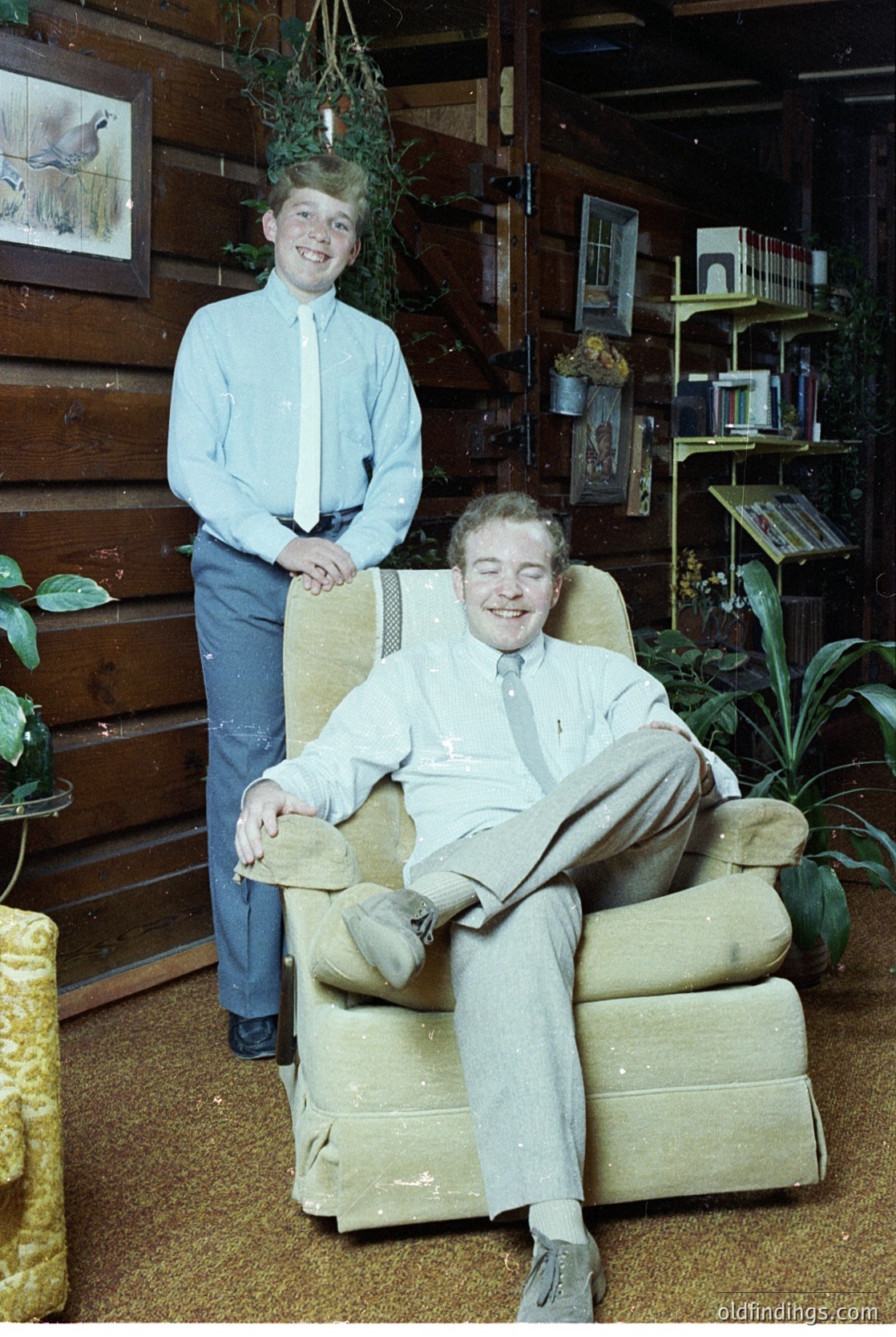 Vintage indoor portrait featuring a seated man in a light-colored suit and tie, relaxed in a vintage armchair, with a standing young boy in a light blue button-up shirt and dark pants beside him. Wood-paneled walls, framed artwork, and potted plants create a mid-century home ambiance.
