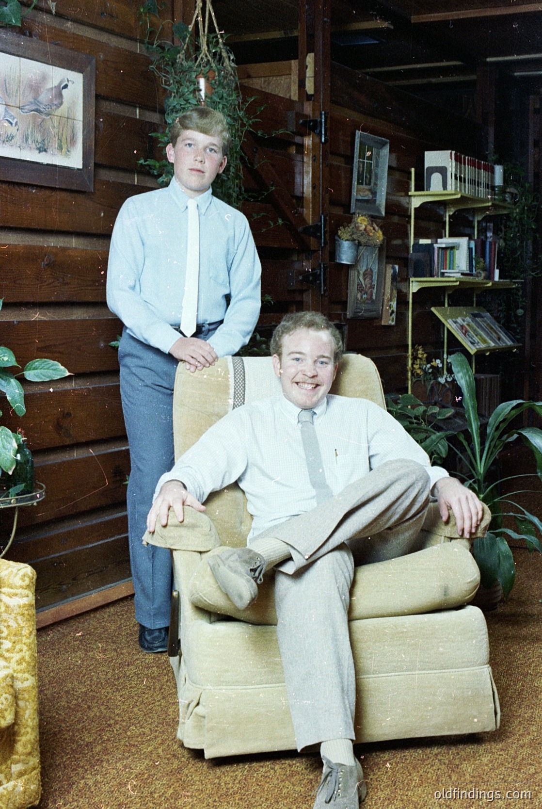 Two men pose indoors in a rustic, book-filled setting, likely mid-20th century. The seated man wears a light-colored suit with a tie, while the standing boy dons a button-down shirt and jeans. Wooden walls, shelves lined with books, and a hanging plant frame the scene. [Mid-20th-century indoor portrait of father and son in a cozy, book-filled home ]