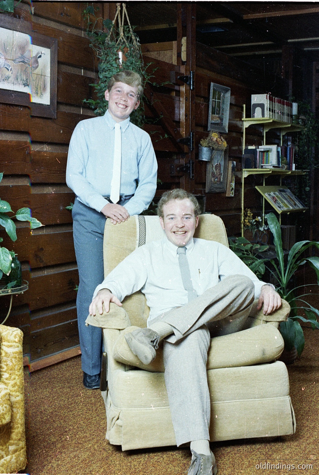 Two men pose indoors in a rustic, book-filled setting, likely mid-20th century (1950s–1960s). The seated man wears a light-colored suit and tie, while the standing man dons a light blue button-up with jeans. Wooden walls, vintage armchair, and hanging plants add to the cozy, scholarly atmosphere.