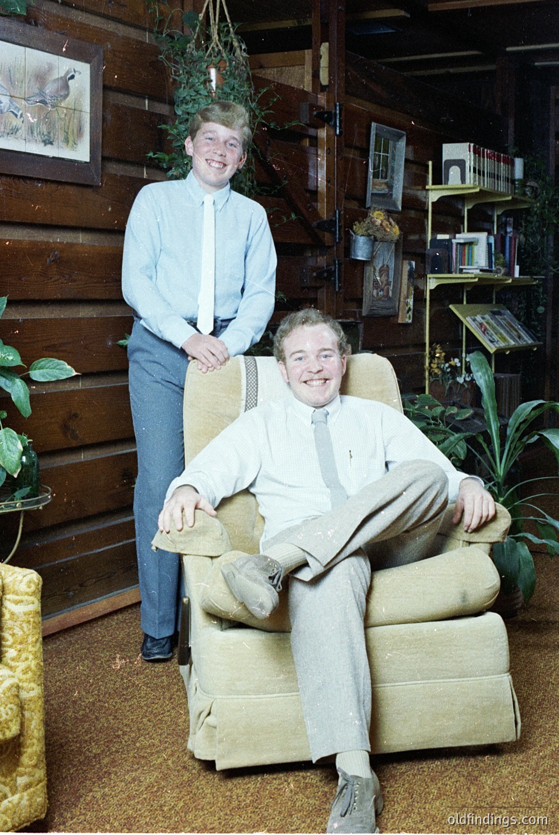 Two individuals pose in a rustic indoor setting, likely mid-20th century (1960s–1970s). The man sits in a vintage armchair, wearing a light vest, dress shirt, and trousers, while the woman stands beside him in a light blue button-down and jeans. Surrounding them: wooden paneling, framed art, potted plants, and shelves with books. Warm, homely atmosphere.