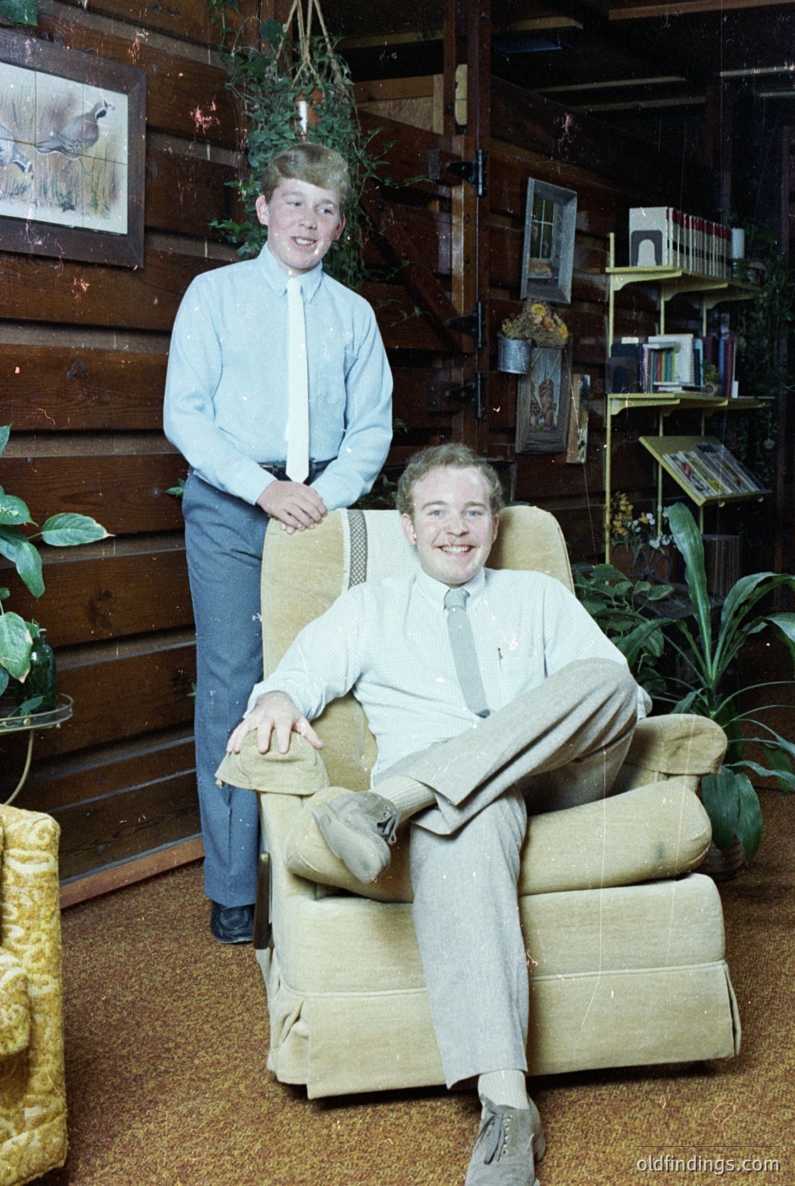 Two men pose indoors in a rustic wooden-paneled room, likely mid-20th century. The seated man wears a light-colored suit with a striped tie, holding a book. The standing man in a light blue dress shirt and dark pants stands behind him. Decor includes vintage bookshelves, hanging plants, and a potted plant on the floor. Warm lighting enhances the cozy, lived-in atmosphere.