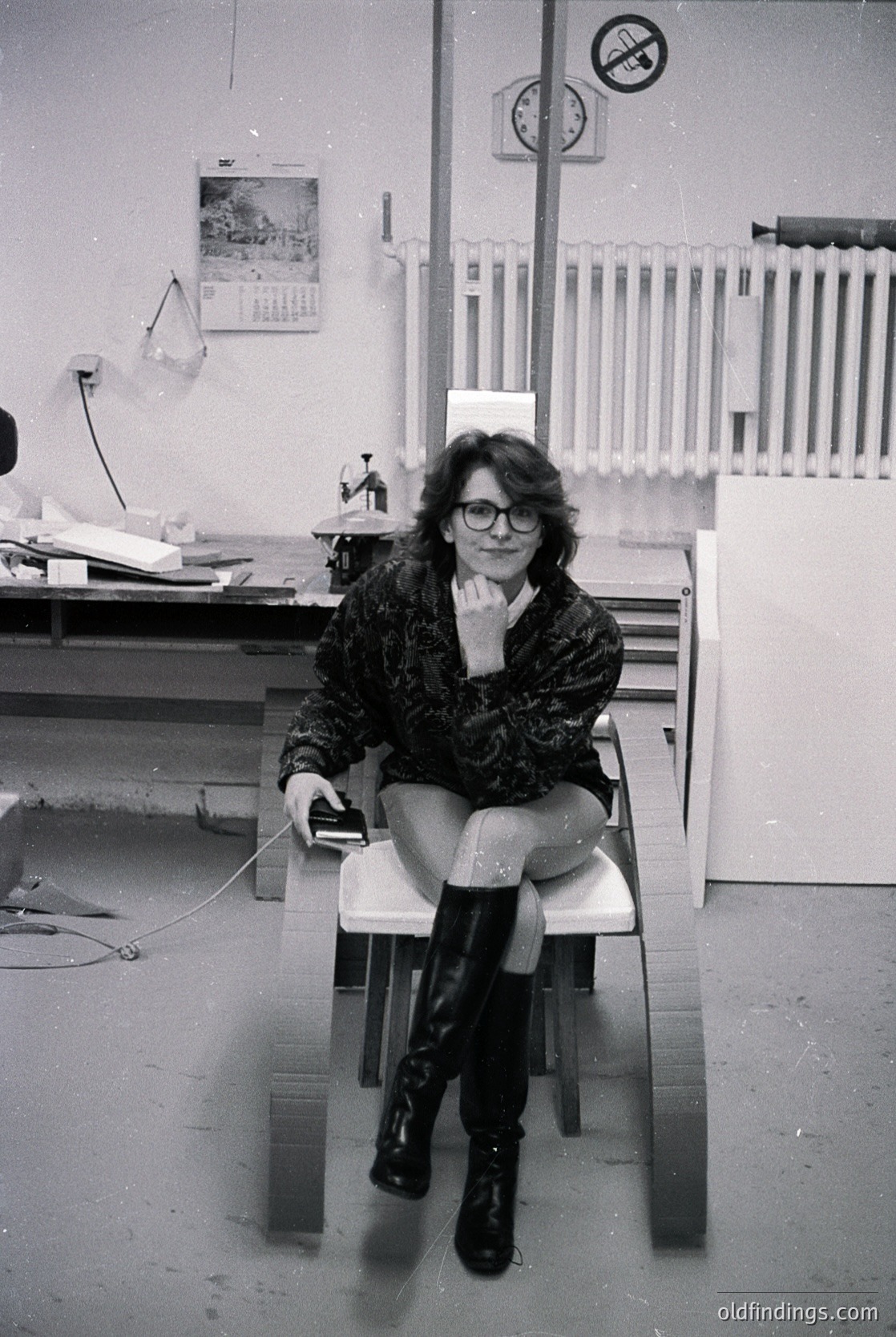 A woman in 1970s-style glasses and patterned coat sits on a white stool in an industrial office, holding a phone receiver. Surrounding elements include a no-smoking sign, a calendar, and a metal radiator. The setting suggests a mid-century workspace.