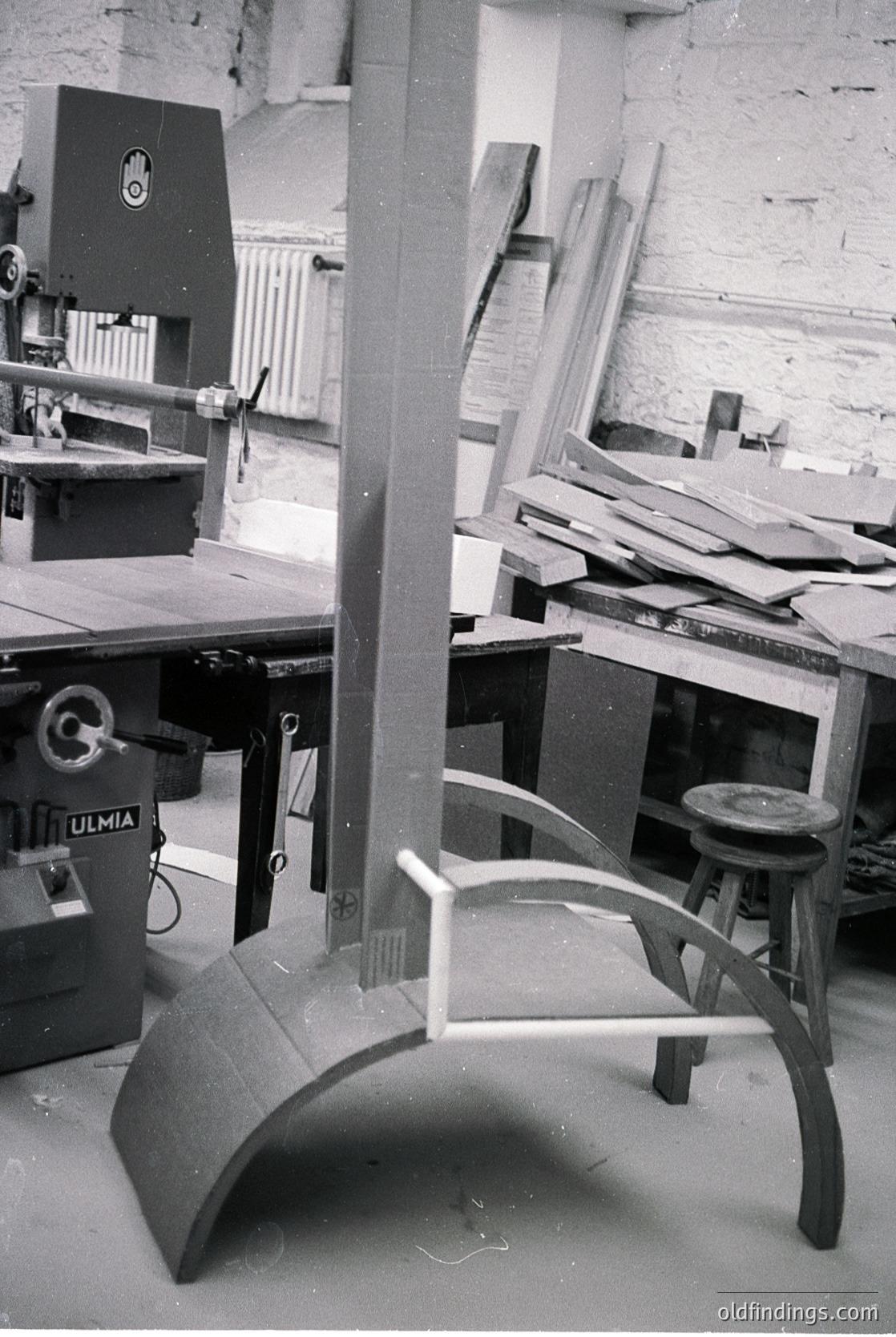 Industrial woodshop featuring vintage machinery: "ULMIA" table saw (left) and a curved wooden bench with curved legs. Stacked plywood sheets and a stool in background. Mid-20th century workshop setting.