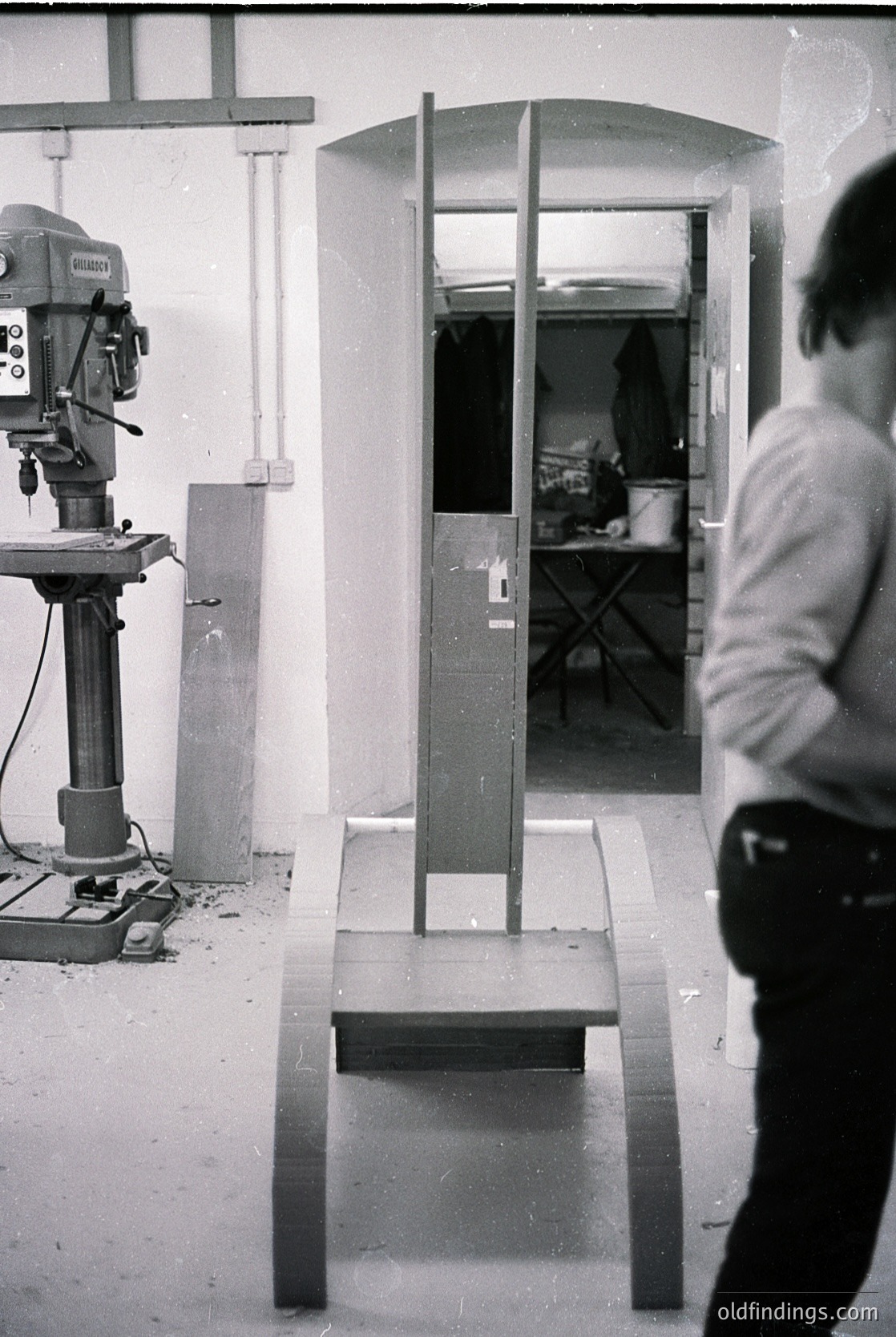Industrial workshop scene featuring a **1960s-70s column saw** (Wirtz brand) with a partially completed metal frame on a workbench. Exposed pipes, a folding chair, and a doorway reveal a utilitarian workspace. Black-and-white grain highlights mid-century craftsmanship.