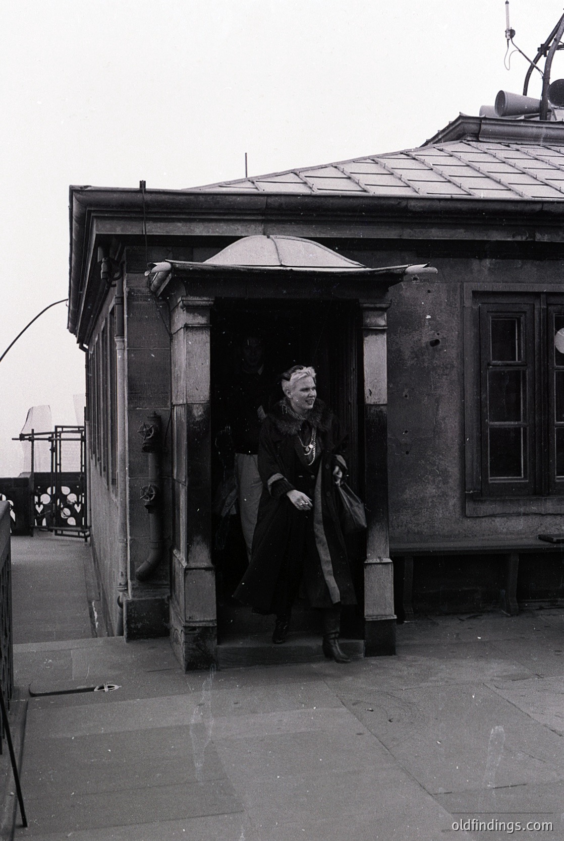 Mid-20th century black-and-white photo of a woman exiting a stone building’s arched entrance, holding a large umbrella and coat. Architectural details include ornate stonework and a sloped roof with visible ventilation pipes. Urban setting with visible street lamps and distant figures.