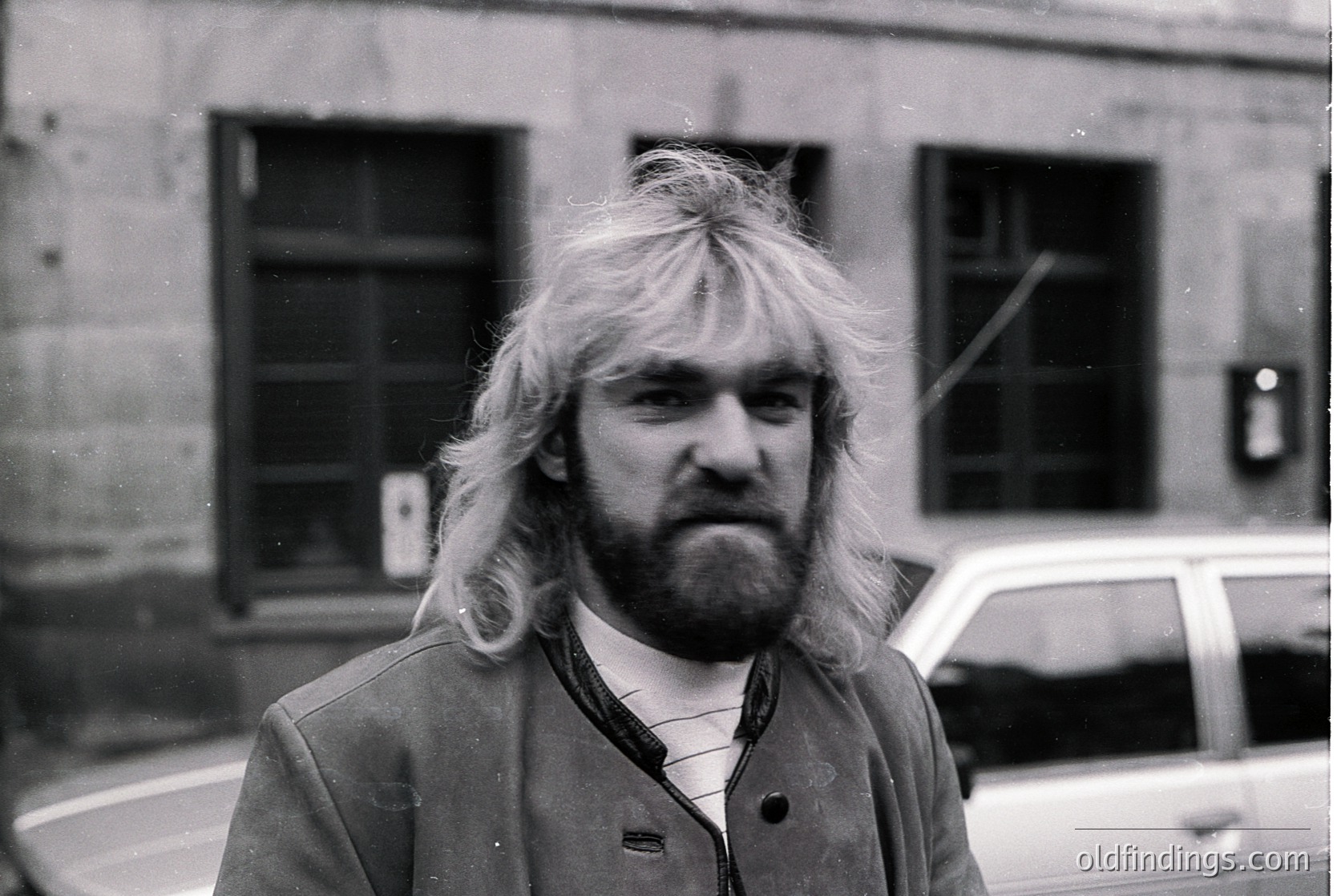 Long-haired man in 1970s urban streetwear—blazer, patterned shirt, and sideburns—posed against a brick building. Vintage car parked in background suggests mid-century European cityscape.