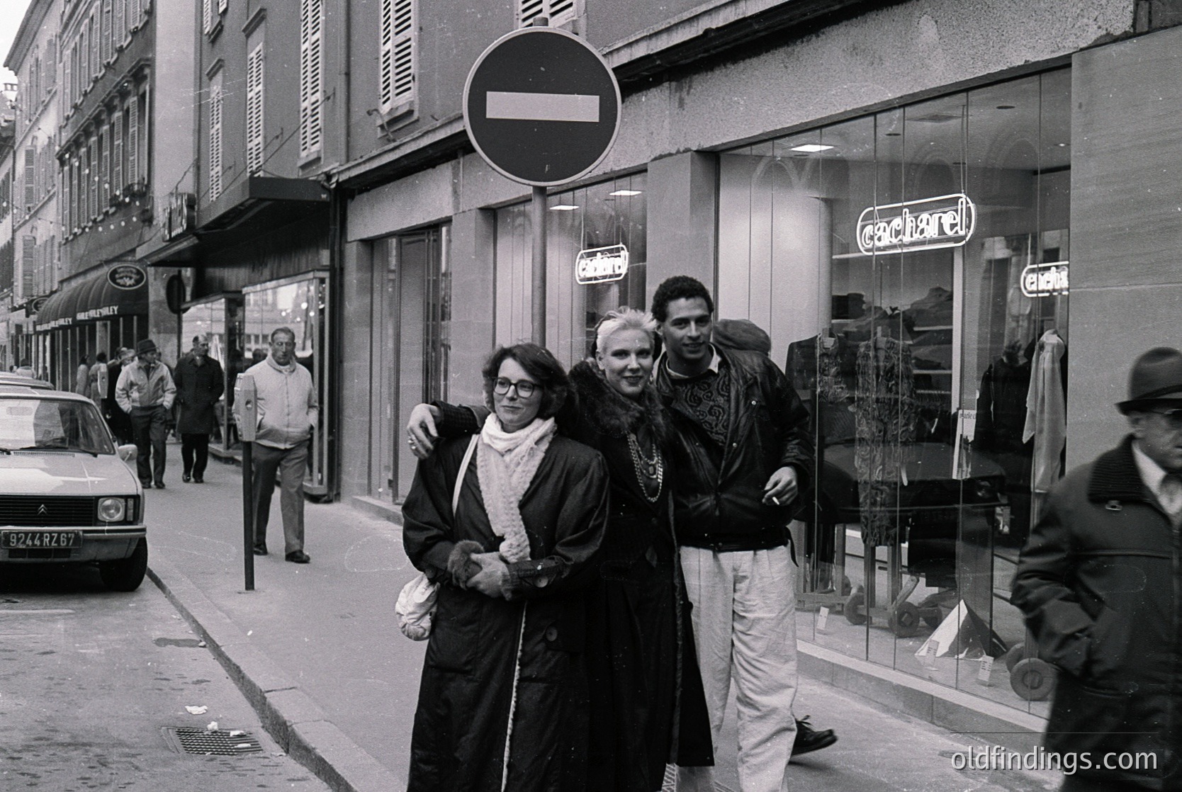 Mid-20th century street scene in Paris, featuring three central figures in 1960s fashion: a woman in a dark coat and scarf, a man in a light sweater, and another in a jacket with a scarf. Surrounding them are classic Parisian storefronts, including a "Cacharel" boutique. A no-entry traffic sign and vintage cars add to the era’s authenticity.
