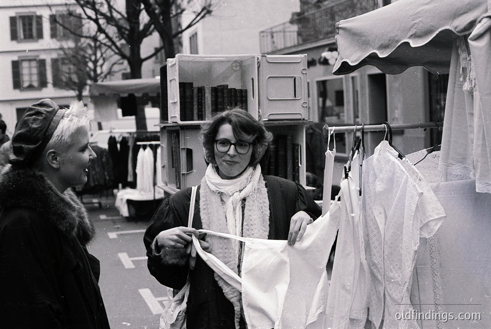 Two women inspecting vintage clothing at an outdoor market stall, likely 1970s–1980s. One holds a sheer blouse; the other wears a fur collar and glasses. Stalls feature draped fabrics and clothing racks. Urban setting with parked vans and residential buildings.