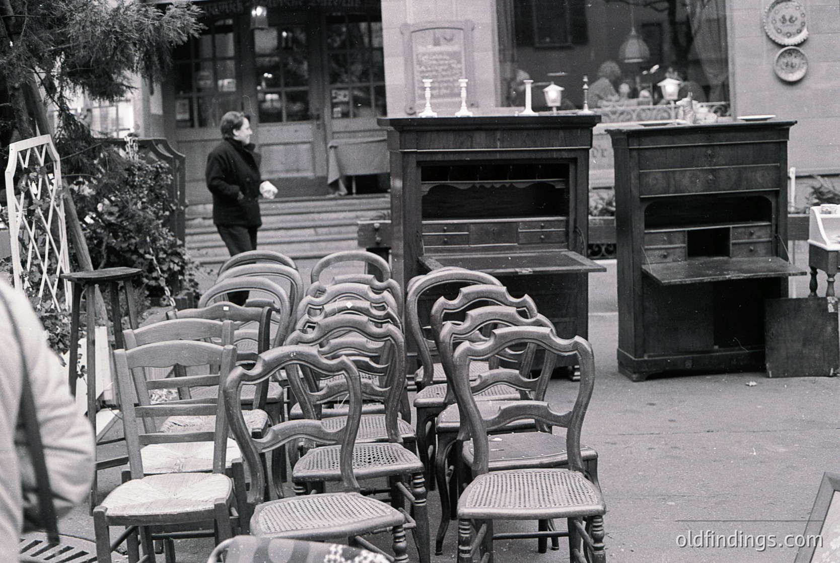 Vintage outdoor café seating with ornate wooden chairs and tables, likely from the 1950s–1970s. Antique display cabinets and a signboard in the background suggest a European street scene. Urban setting with classic mid-century architecture. é