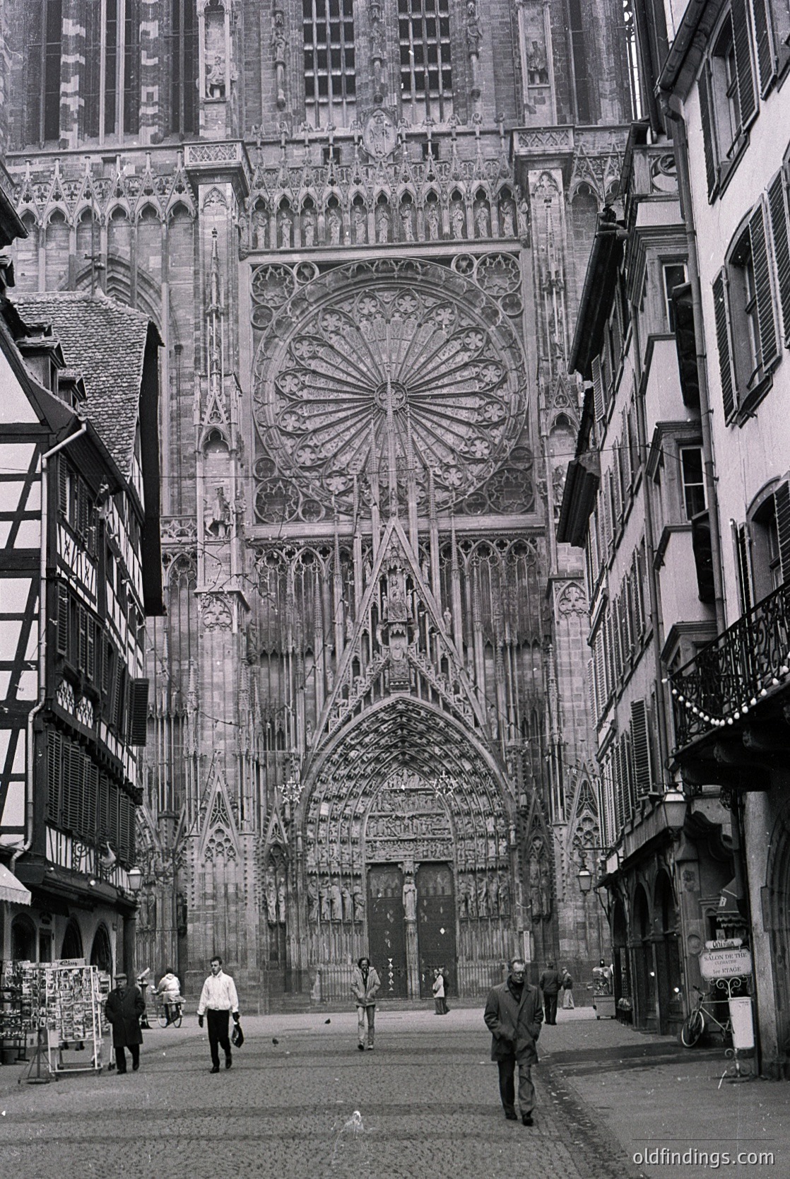 Gothic façade of Strasbourg Cathedral’s south portal, flanked by half-timbered medieval buildings. Intricate stone carvings, rose window, and pointed arches dominate the 13th–15th century façade. Cobblestone street with pedestrians in 1960s-era clothing.