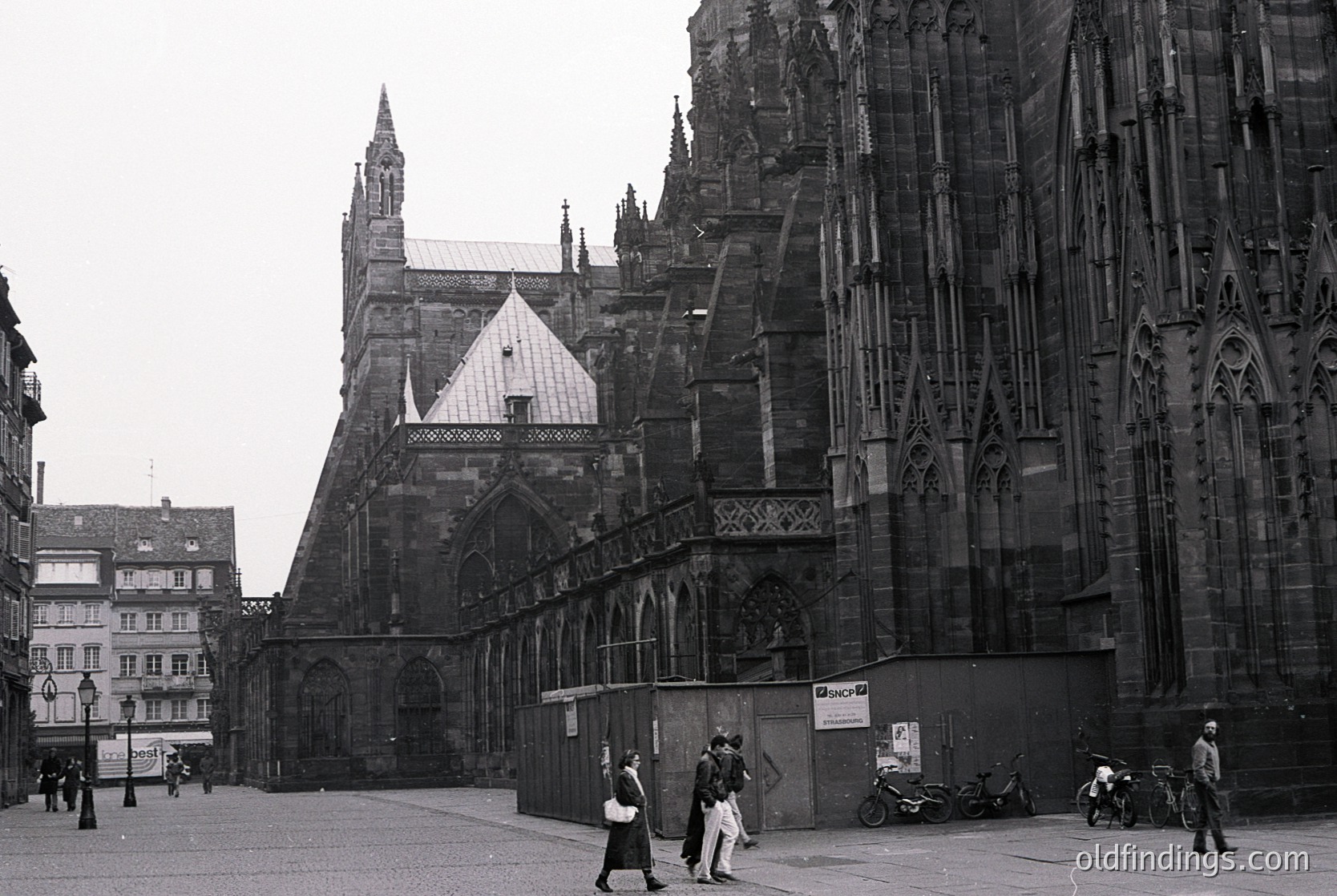 Gothic cathedral façade with intricate stonework and pointed arches, likely **Ulrichskirche (St. Ulrich’s Church)** in **Augsburg, Germany**. Black-and-white photo captures 1950s–1960s urban life with pedestrians and motorcycles. Architectural details include ornate tracery and a steeply pitched roof.