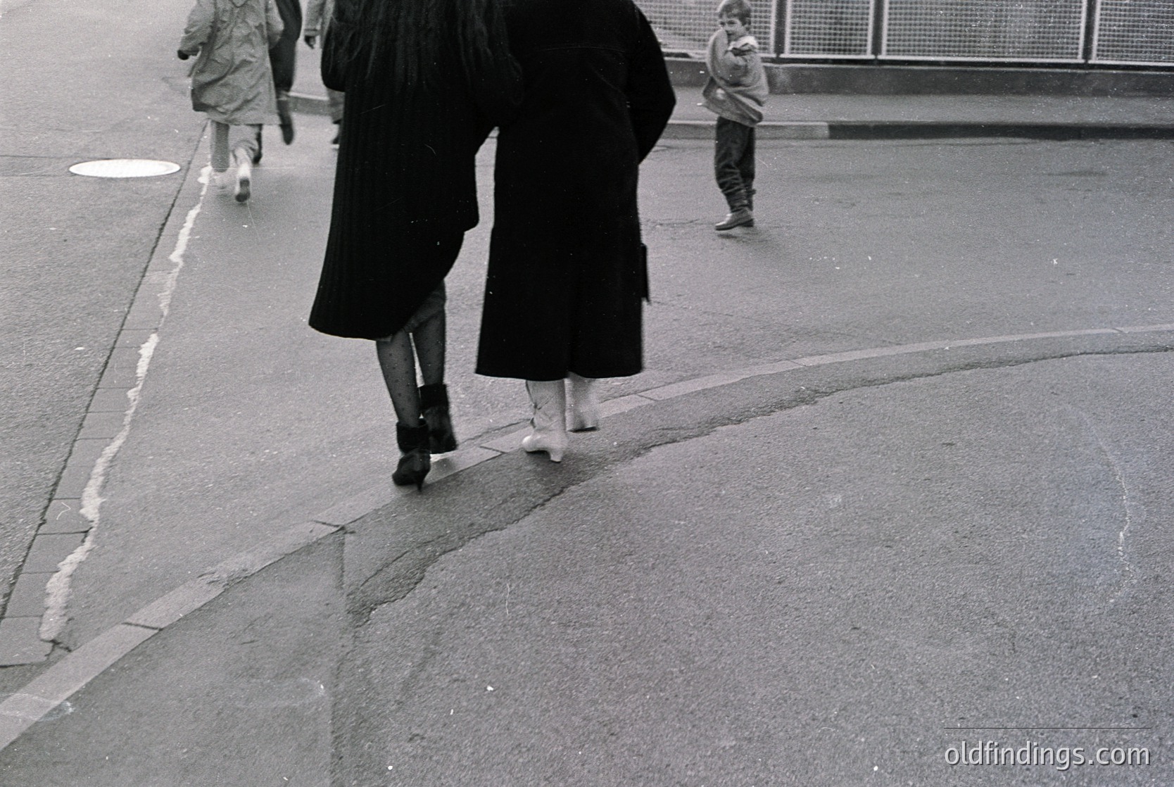 Black-and-white street scene featuring two women in long, floor-length coats and ankle boots crossing a curved sidewalk. One woman wears a wide-brimmed hat; the other’s coat drapes over a child’s head. Background shows indistinct pedestrians and a metal railing. Mid-century urban design, likely 1950s–1960s.