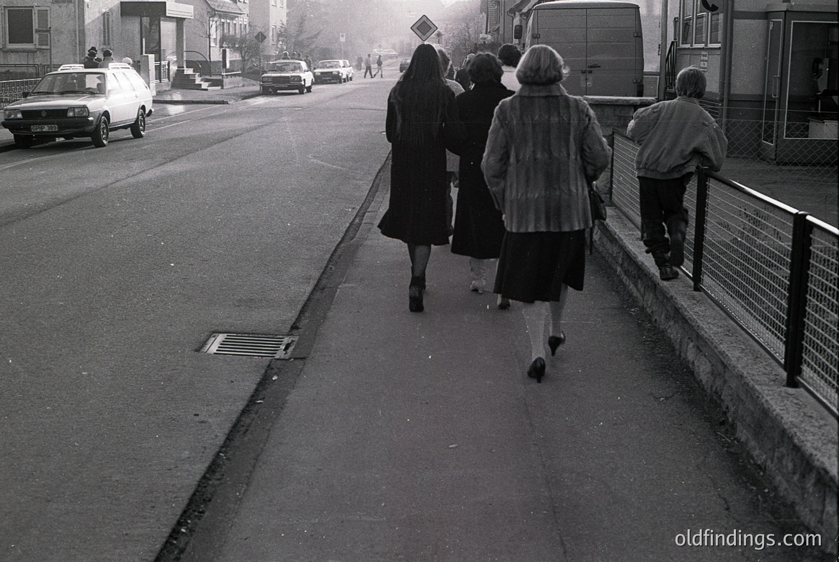 Four individuals in 1970s-era attire walk away from the camera on a paved sidewalk beside a road. The woman in the center wears a dark coat and knee-length skirt; others in coats and scarves. Vehicles, including a white van and sedan, share the road with a pedestrian crossing sign. Urban setting with residential buildings in the background.