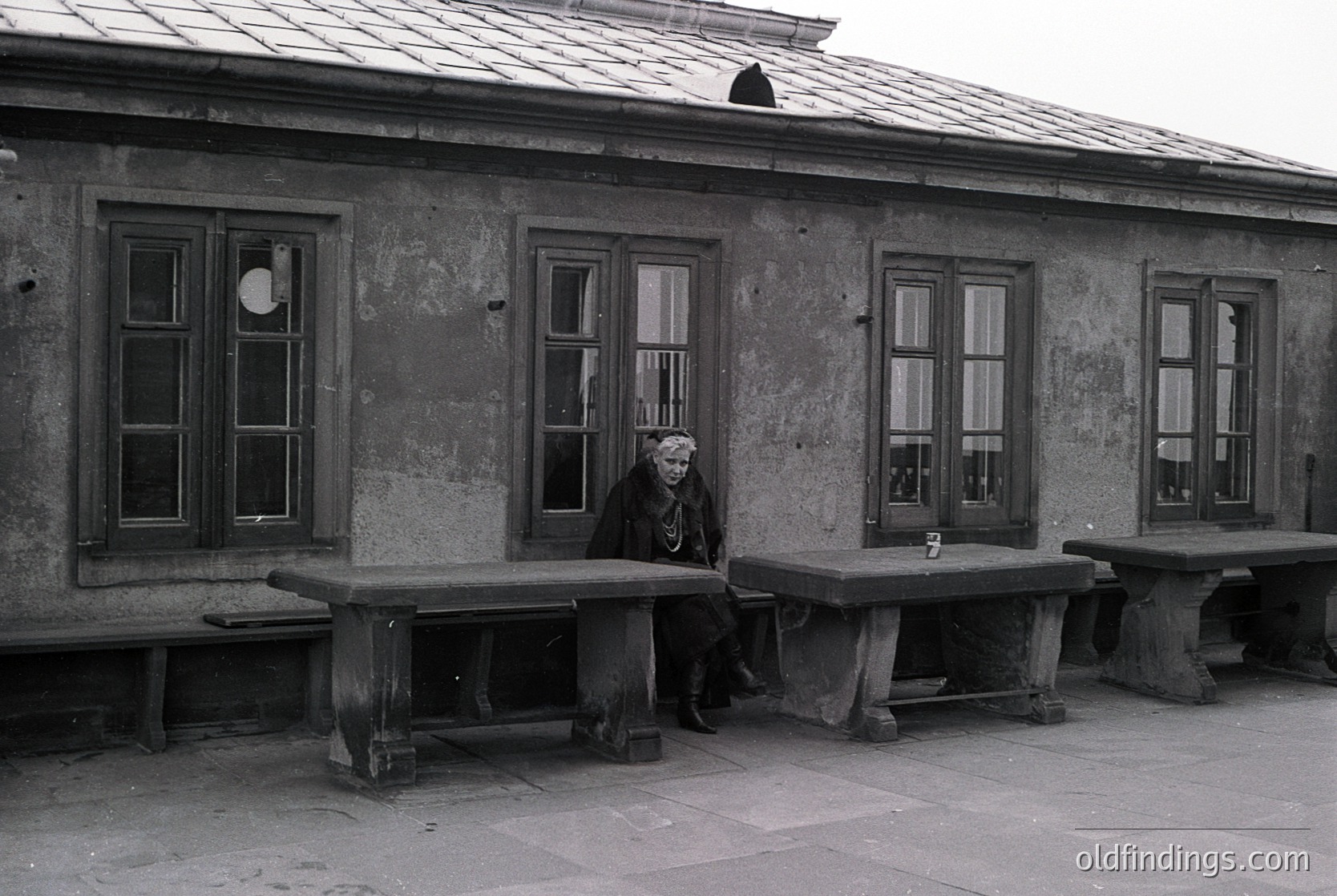 Black-and-white photo of elderly woman seated on concrete bench outside weathered concrete building with large rectangular windows. Simple wooden table beside her. Likely Eastern European institutional architecture, 1970s–1980s.