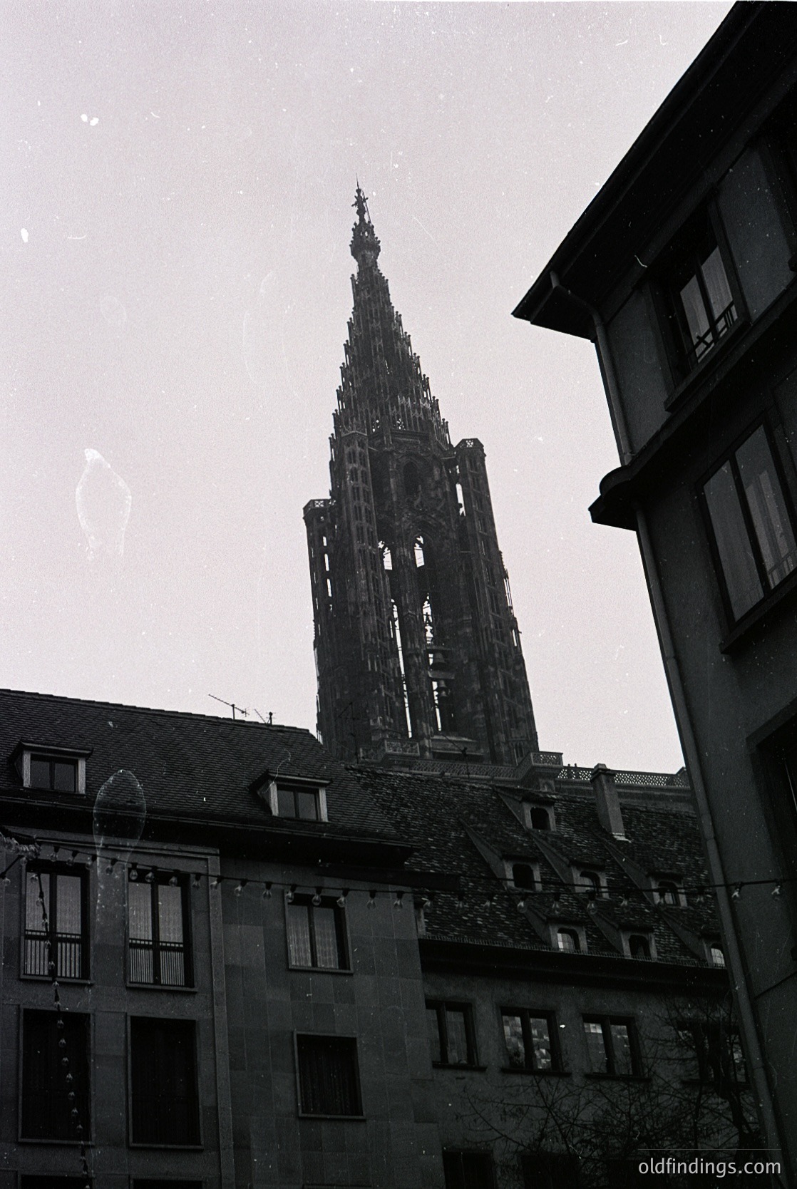 Gothic spire dominates urban skyline, framed by adjacent residential buildings with slate roofs and wrought-iron balconies. Likely Strasbourg Cathedral, 12th–15th century, exemplifying French Gothic architecture.