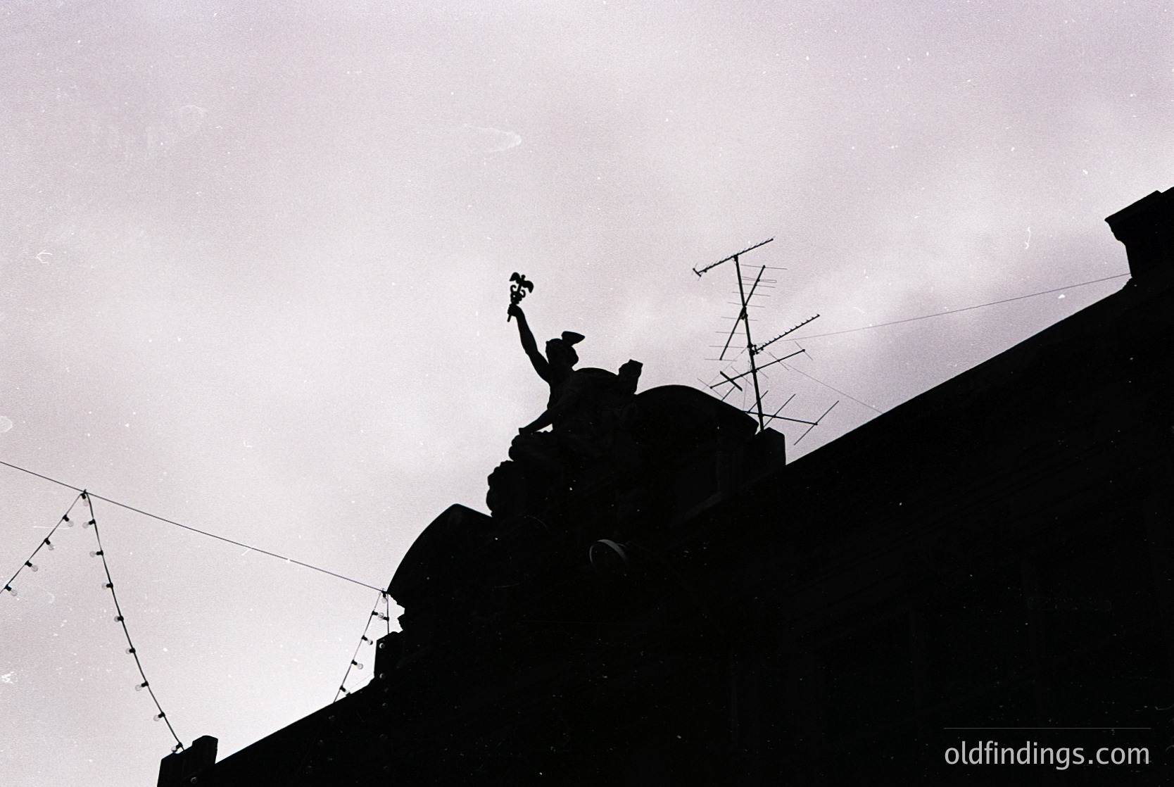 Silhouetted figure atop a rooftop, raising a small object (likely a flag or flower) against a cloudy sky. Urban setting with visible barbed wire fencing and antennae. Suggests a moment of protest or celebration. Black-and-white, high-contrast style indicative of mid-20th century photography.