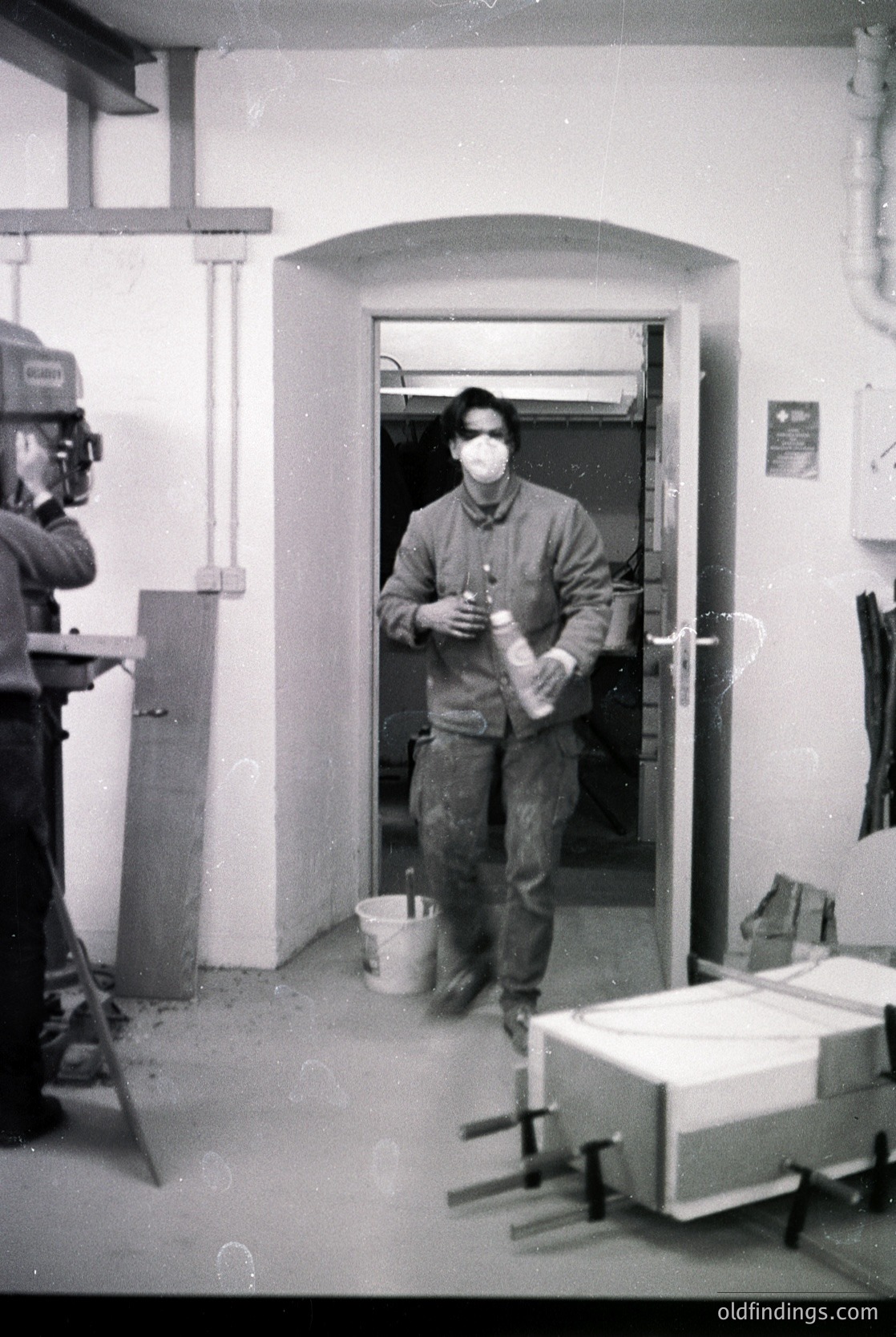 Black-and-white industrial scene: man in 1970s workwear (overalls, mask) exits through an arched doorway holding a bucket, with a film camera crew filming. Equipment includes a tripod-mounted camera and a dolly. Plain concrete walls and floor suggest a factory or construction site.