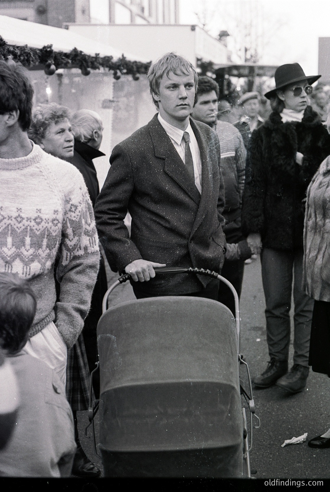 Mid-20th century street scene featuring a man in a suit and tie pushing a baby buggy. Crowd in winter attire—knit sweaters, coats, and hats—suggests cold weather. Urban setting with blurred background indicating movement. Likely 1960s–1970s, Western Europe or North America.
