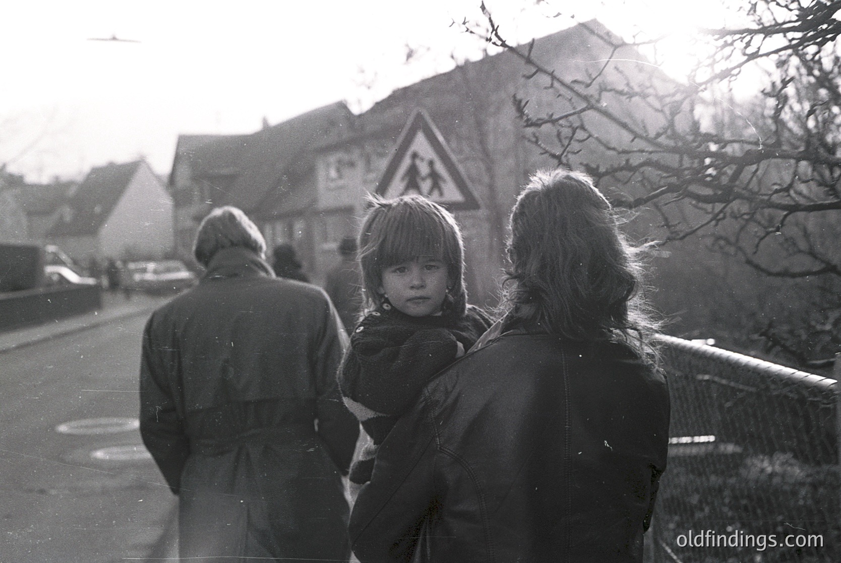 Black-and-white street scene featuring three adults walking with a child in a rural or small-town setting. The adult holding the child wears a dark coat and carries a bag; others wear layered clothing. A triangular pedestrian warning sign and traditional European-style houses with steep roofs frame the background. Likely 1970s–1980s based on clothing and signage.