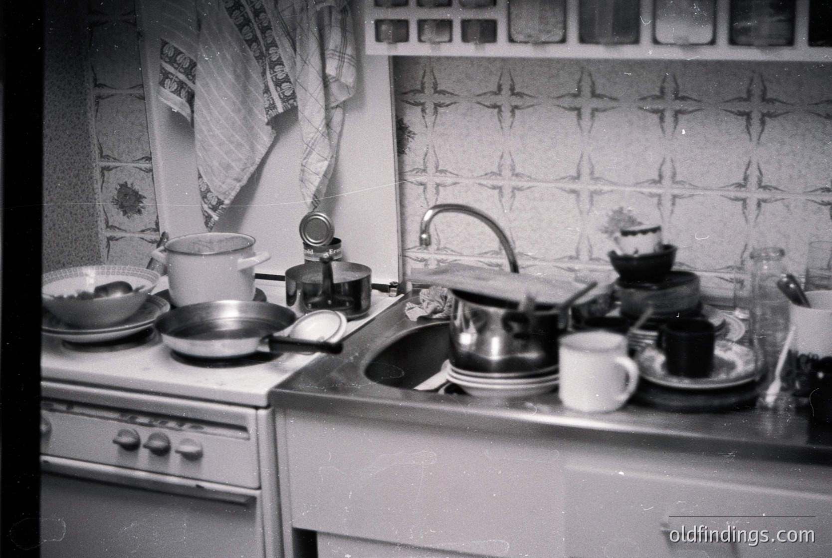 Mid-century kitchen with vintage stove, sink, and countertop cluttered with dishes, a teapot, and a coffee maker. Geometric wallpaper features star motifs. Likely Eastern Bloc design, 1960s–1970s.