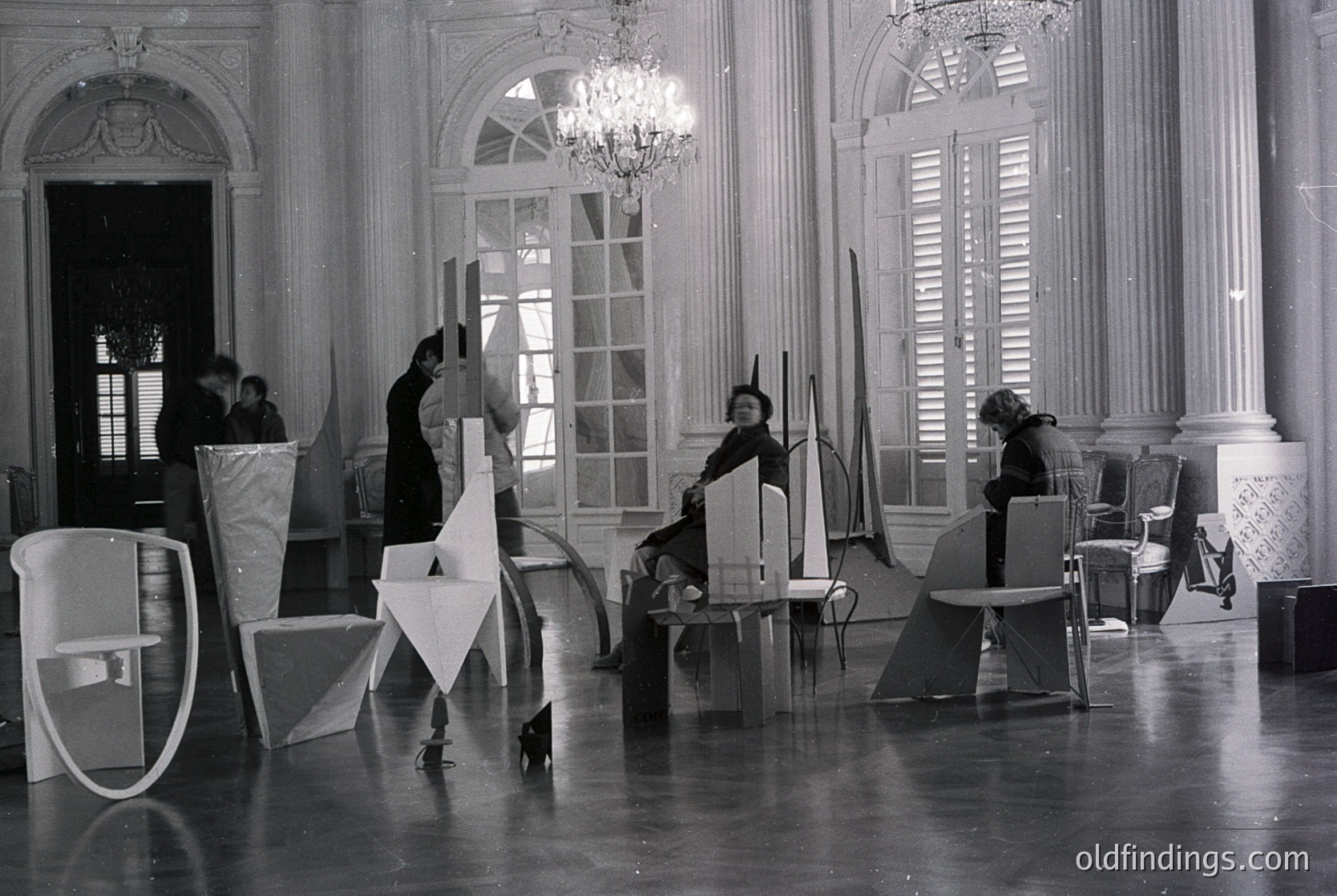 Mid-century modern furniture design studio in a grand, classical interior. Four individuals collaborate with geometric, minimalist chairs and tables, likely from the 1960s–1970s. High ceilings, arched windows, and ornate moldings contrast with the clean lines of the furniture.