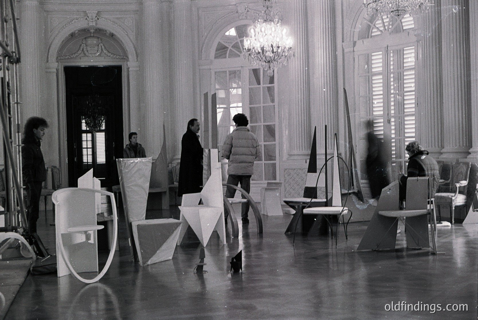 Elegant 1960s–70s interior featuring geometric modernist furniture and grand classical architecture. White plastic chairs and abstract forms contrast with ornate arches, chandeliers, and large windows. Likely a design exhibition or architectural study.