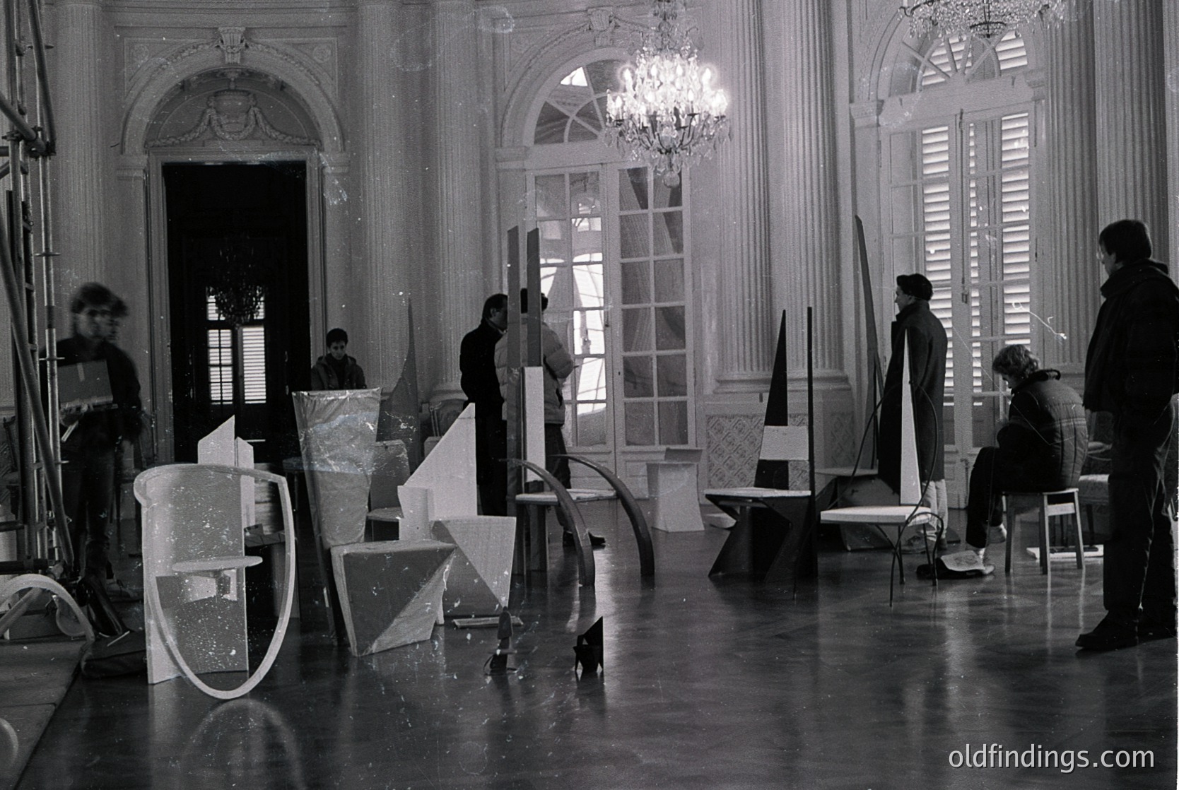 Black-and-white shot of a grand interior featuring geometric set pieces and crew positioning in a spacious, ornate room. Ornate moldings, arched doorways, and large windows with shutters frame the scene. Likely a 1960s–1970s film set, possibly European due to architectural style.