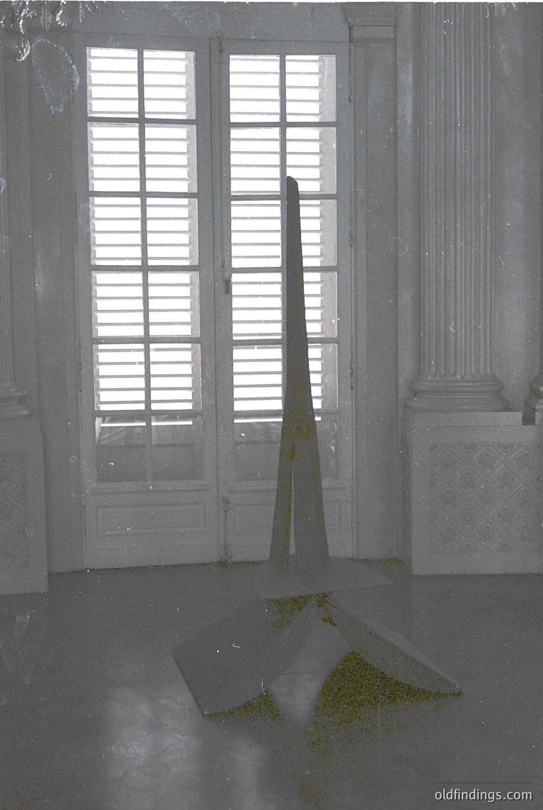 Neoclassical interior featuring tall, open double doors with louvered upper panels and frosted glass lower sections. A single, narrow, triangular skylight casts angled light onto a concrete floor with moss growth. Ornate plaster corbels and a partial column frame the doorway. Likely a historic European building, possibly 19th–early 20th century. #