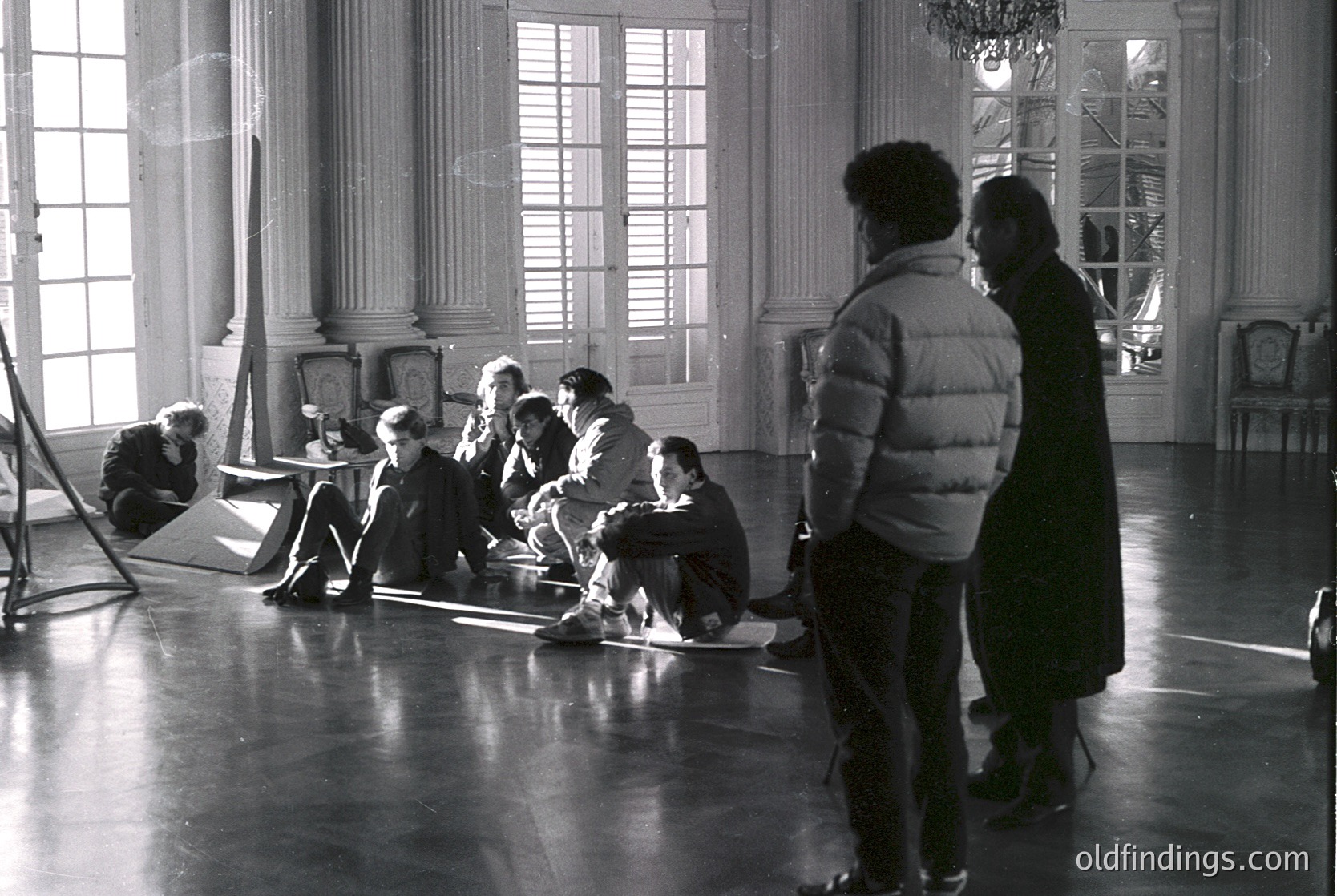 Grand ballroom setting with classical architecture: ornate columns, high ceilings, and large arched windows. Group of people in 1970s attire—men in suits, women in dresses and jackets—engaged in discussion. One person kneels, others sit on floor. Lighting suggests indoor filming or rehearsal.