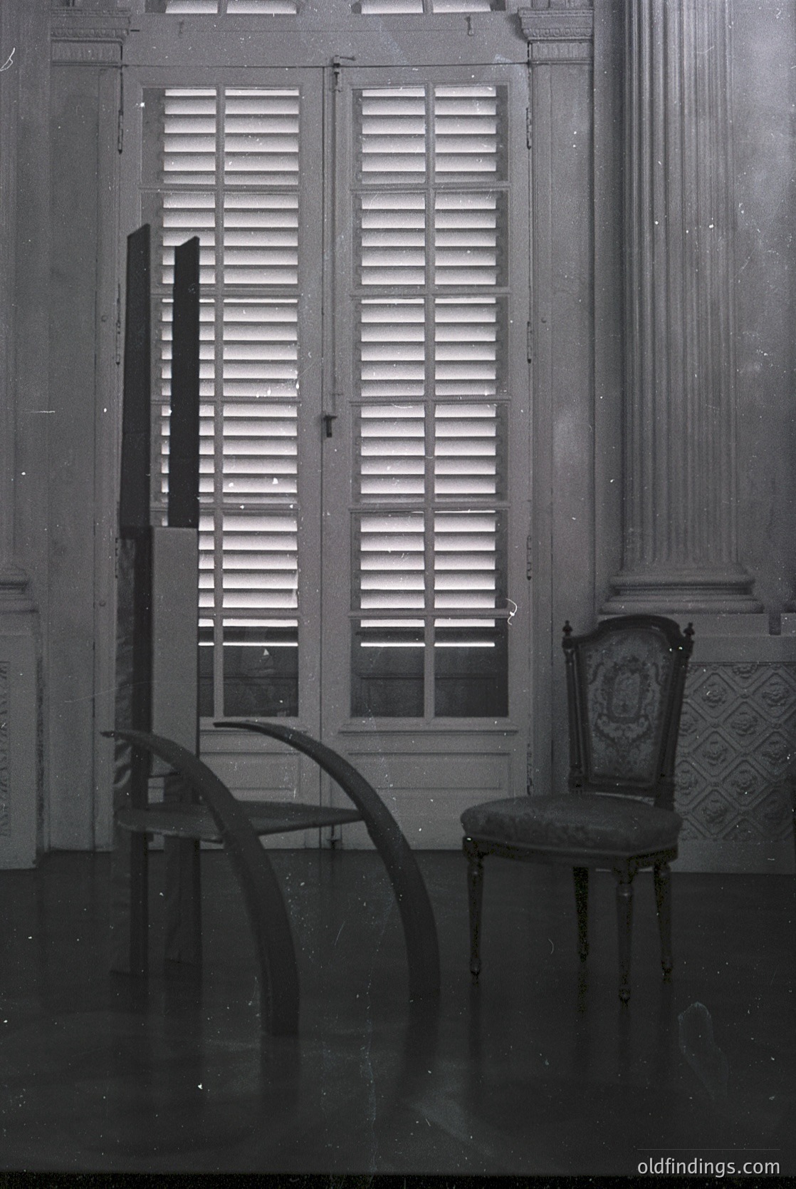 Vintage interior featuring classic French-inspired design: closed louvered shutters casting geometric light patterns, a curved wooden armchair with ornate carvings, and a tufted upholstered chair beside a marble column. Likely mid-20th century European residential architecture.