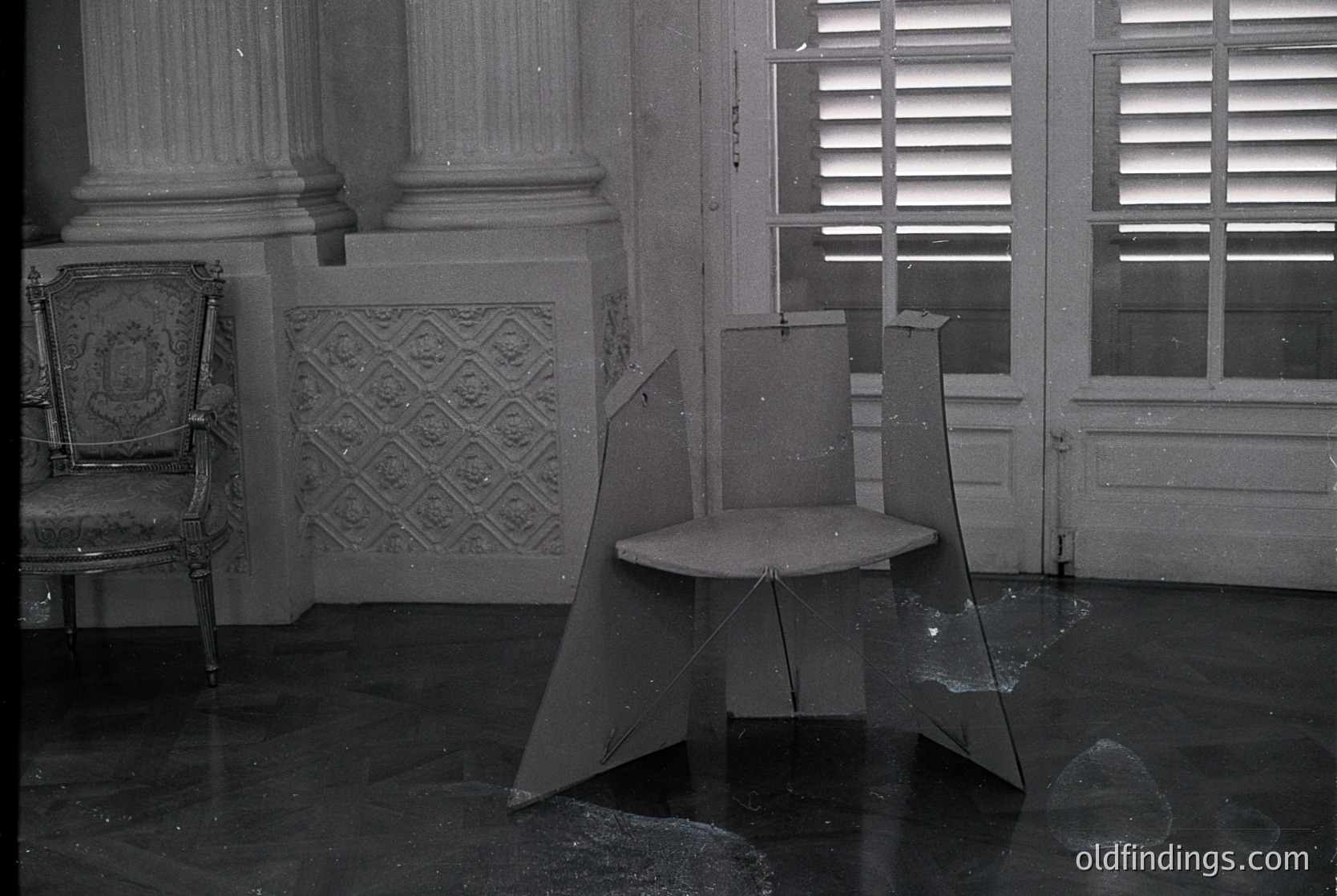 Vintage black-and-white interior of a grand, abandoned room with ornate plasterwork and shuttered windows. Broken glass and a collapsed stool in foreground; upholstered armchair with intricate detailing in background. Likely mid-20th century European residential or institutional architecture.