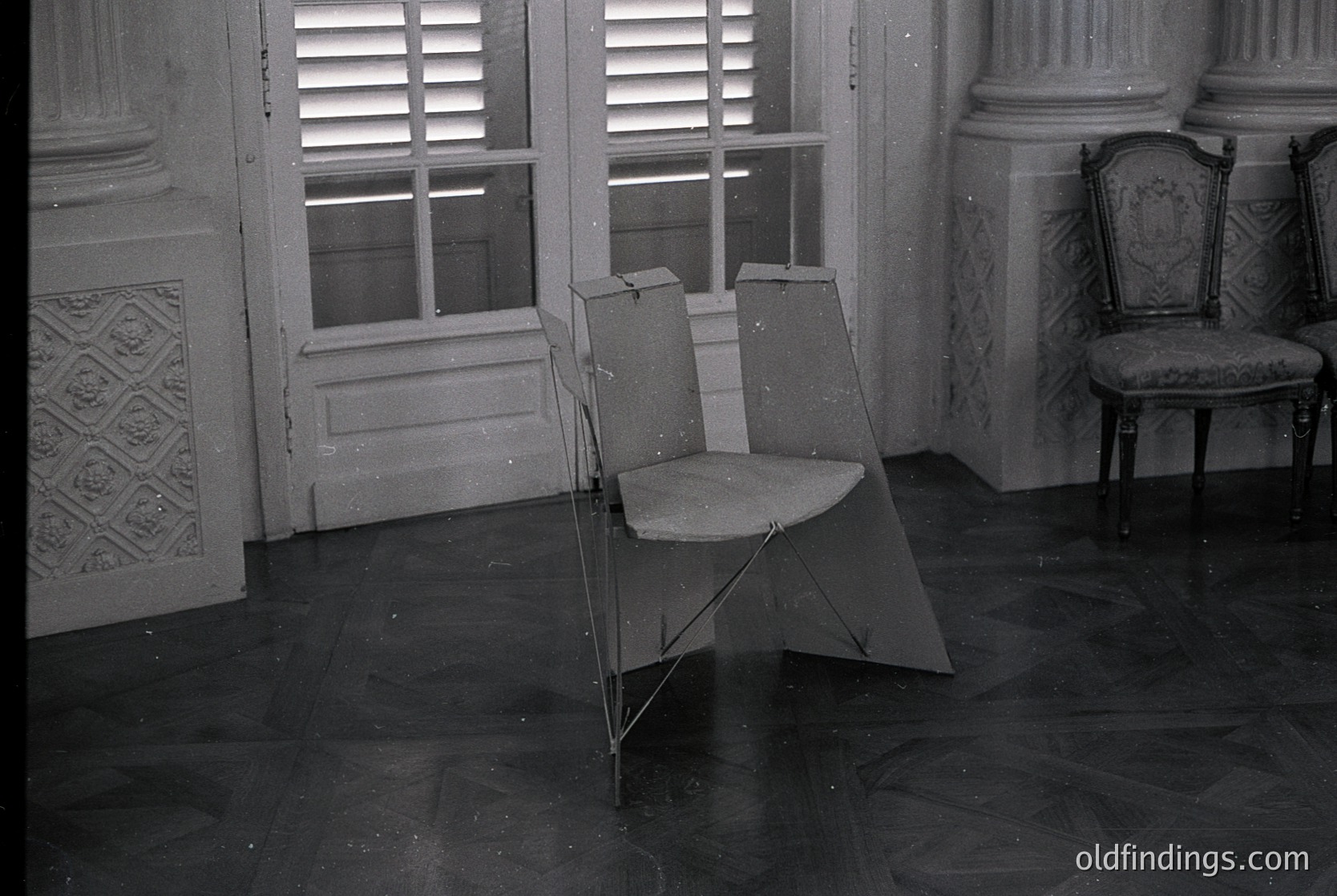 Vintage folding chair placed beside an ornate, upholstered armchair in a grand, dimly lit room. Decorative plasterwork and shuttered windows suggest classical European interior design, likely from the early-to-mid 20th century. Dark, polished floor contrasts with aged wall textures.