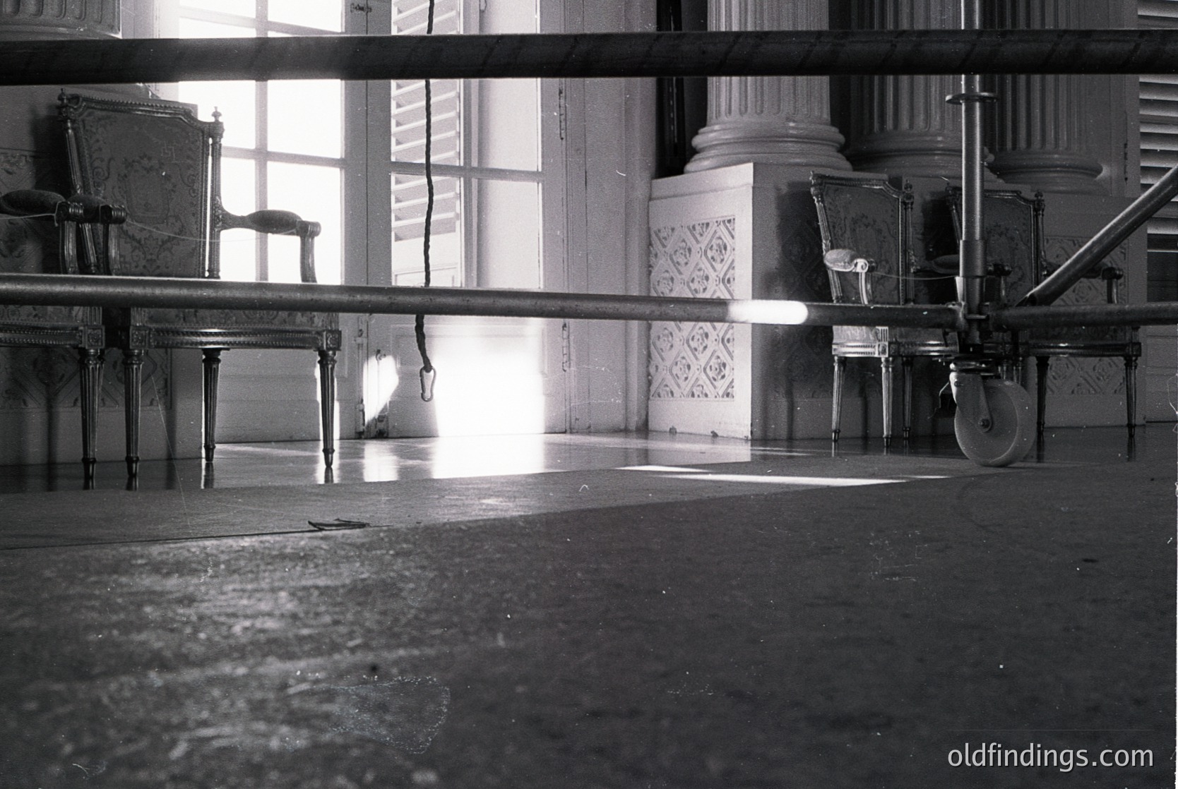 Vintage black-and-white shot of a grand, empty ballroom with classical architecture—ornate plasterwork, high ceilings, and large windows. A vintage projector on wheels sits center-stage, flanked by ornate chairs. Sunlight streams through windows, casting dramatic shadows. Likely 1940s–1960s European venue.