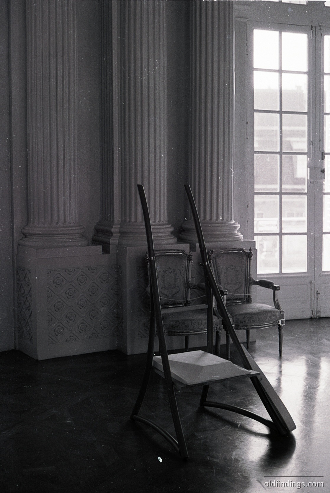 Neoclassical interior featuring tall fluted columns with ornate capitals. Two vintage chairs with curved wooden frames and patterned upholstery placed beside a ladder against a polished marble floor. Large arched windows with divided panes allow natural light to flood the space. Likely a historic European residence or public building, 19th–early 20th century.