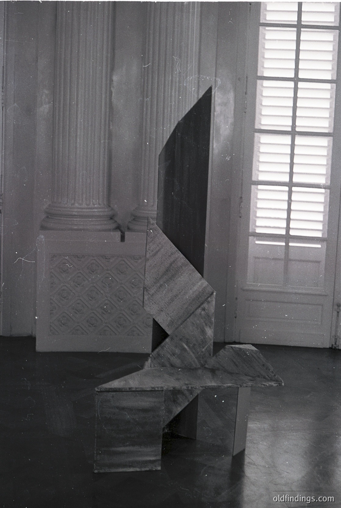 Neoclassical interior featuring geometric marble sculpture and architectural details. The angular, abstract bench contrasts with fluted Corinthian columns and patterned wall panels. Natural light filters through tall, divided windows. Likely 19th–early 20th century European.