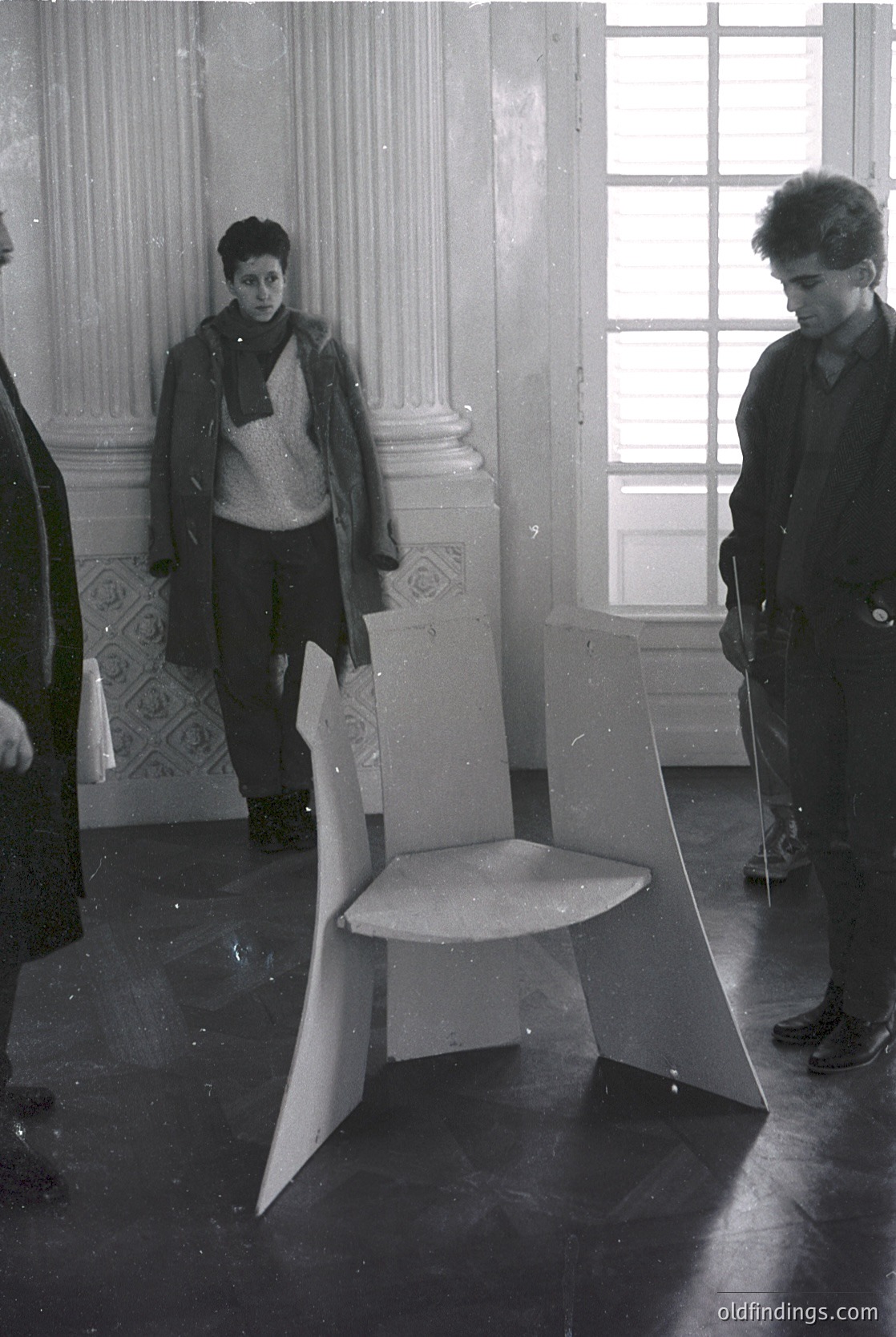 Vintage black-and-white photo of two individuals examining a modernist, angular stool in a grand interior. Ornate plasterwork and large windows suggest a historic European setting, likely mid-20th century. The geometric design contrasts with classical architecture, hinting at avant-garde art or design movements.