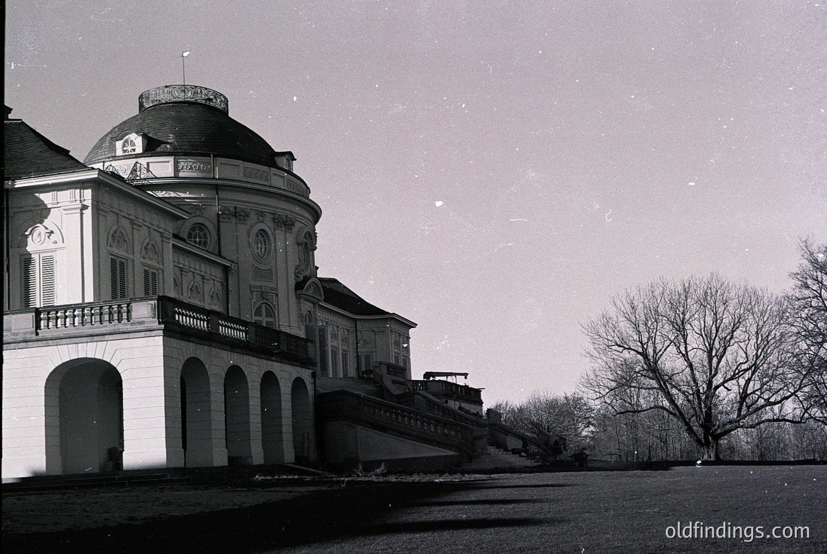Neoclassical villa with domed roof and arched colonnades, captured in monochrome. Ornate stonework and balustrades highlight its 18th–19th century European design. Snow-dusted ground and bare trees suggest winter, likely Central/Eastern Europe.