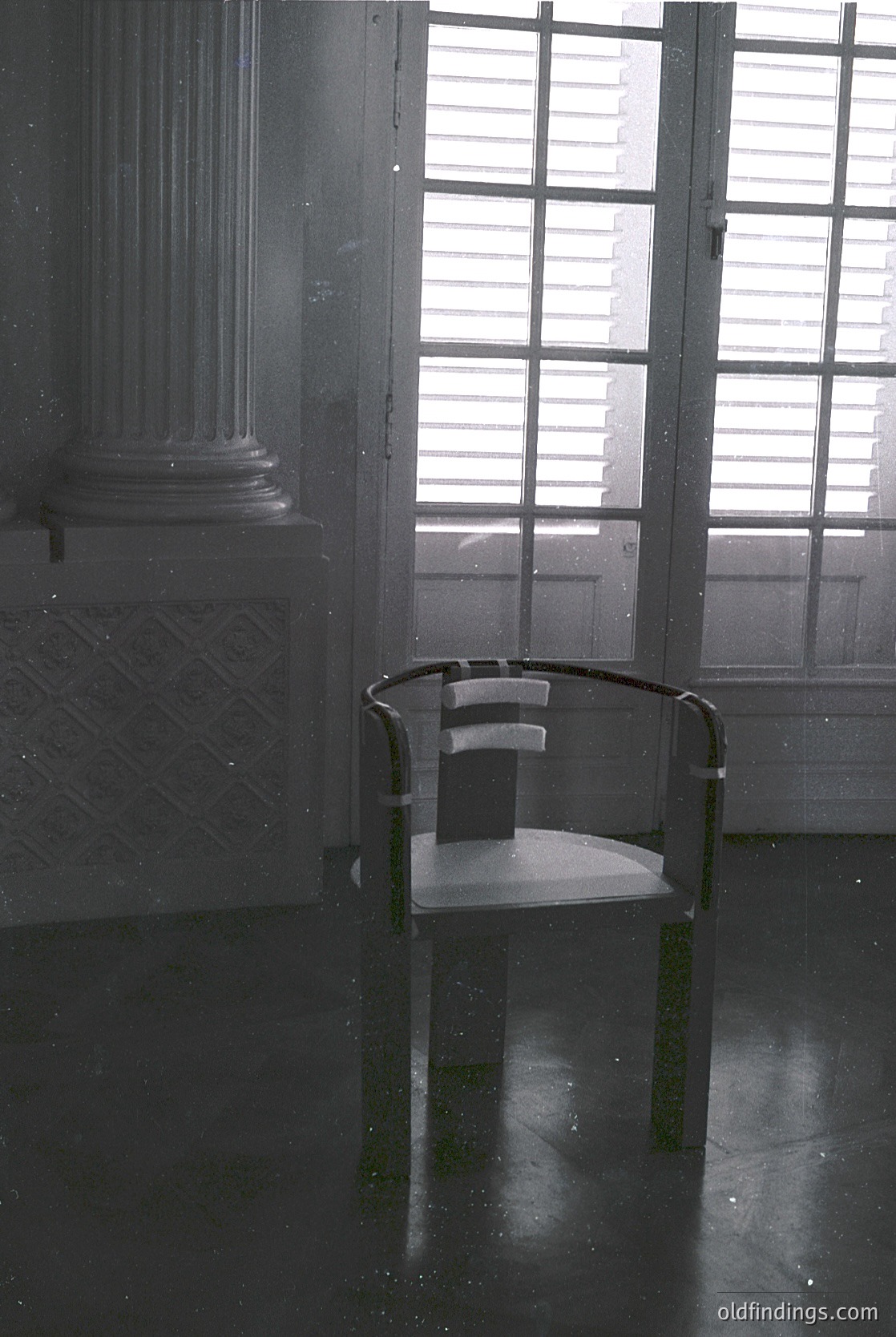 Minimalist interior featuring a single modernist chair with black metal frame and white seat, positioned in a dimly lit room with classical architectural elements. Large shuttered window allows diffused natural light. Geometric floor tiles and a fluted column suggest mid-century design. Ideal for interior design inspiration or historical architecture studies.