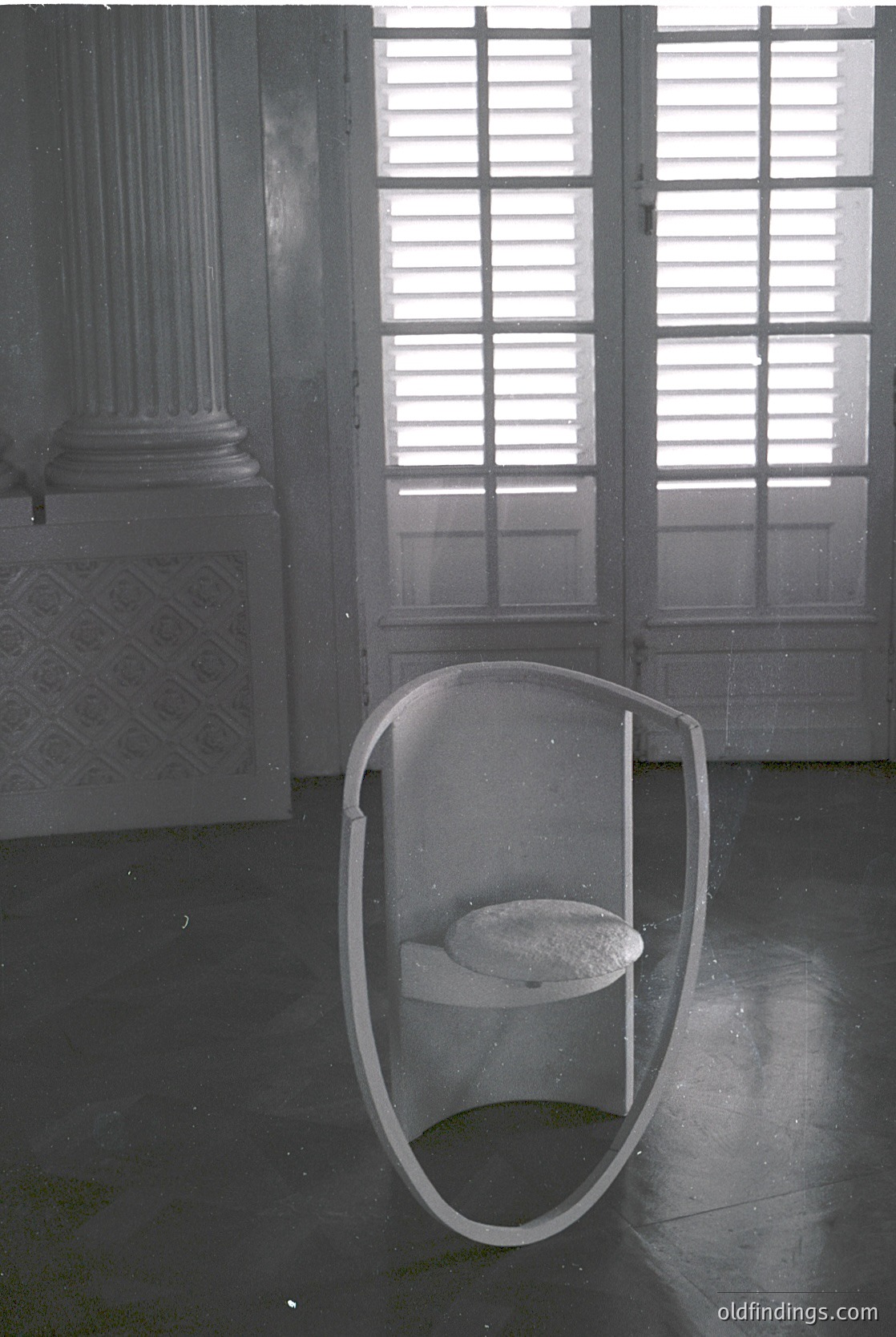Mid-century modern urinal with transparent acrylic bowl, mounted on a tiled floor in a grand, classical-style interior. Fluted columns and louvered doors suggest a public or institutional setting, likely from the 1950s–1970s.