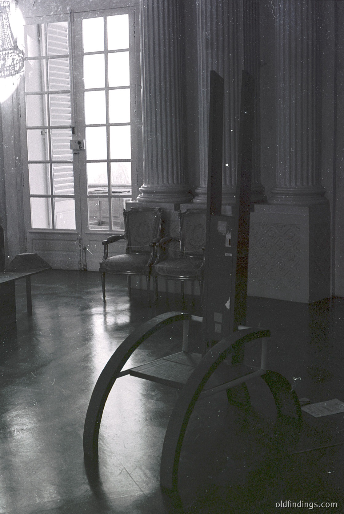 Neoclassical interior featuring grand double doors with frosted glass panes, flanked by fluted Corinthian columns. A vintage armchair with ornate carvings sits beside a polished marble floor. A curved, wrought-iron table with green moss adds rustic contrast. Natural light floods the space, highlighting architectural details. Likely 19th-century European palace or mansion.