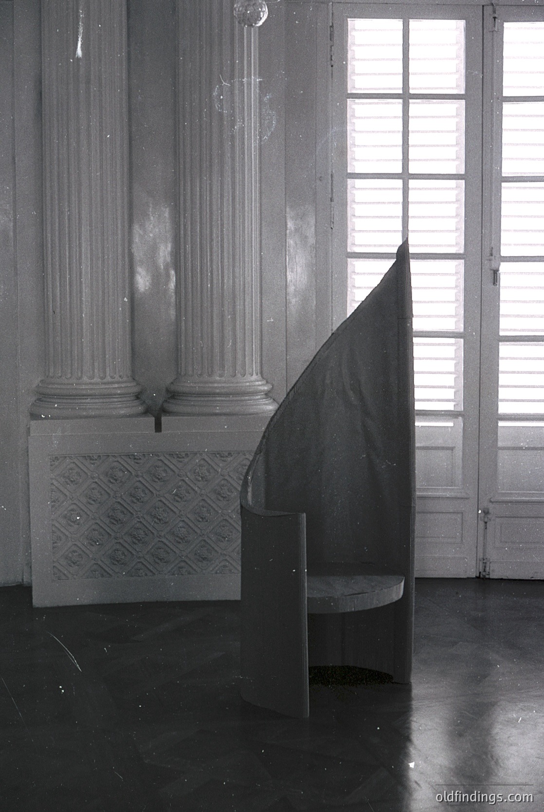 Neoclassical interior featuring two fluted Corinthian columns with decorative capitals flanking a marble fireplace. A draped fabric shadow obscures the hearth, while natural light streams through tall, divided paned windows. Polished marble floor reflects subtle architectural details. Likely European, 19th–early 20th century.