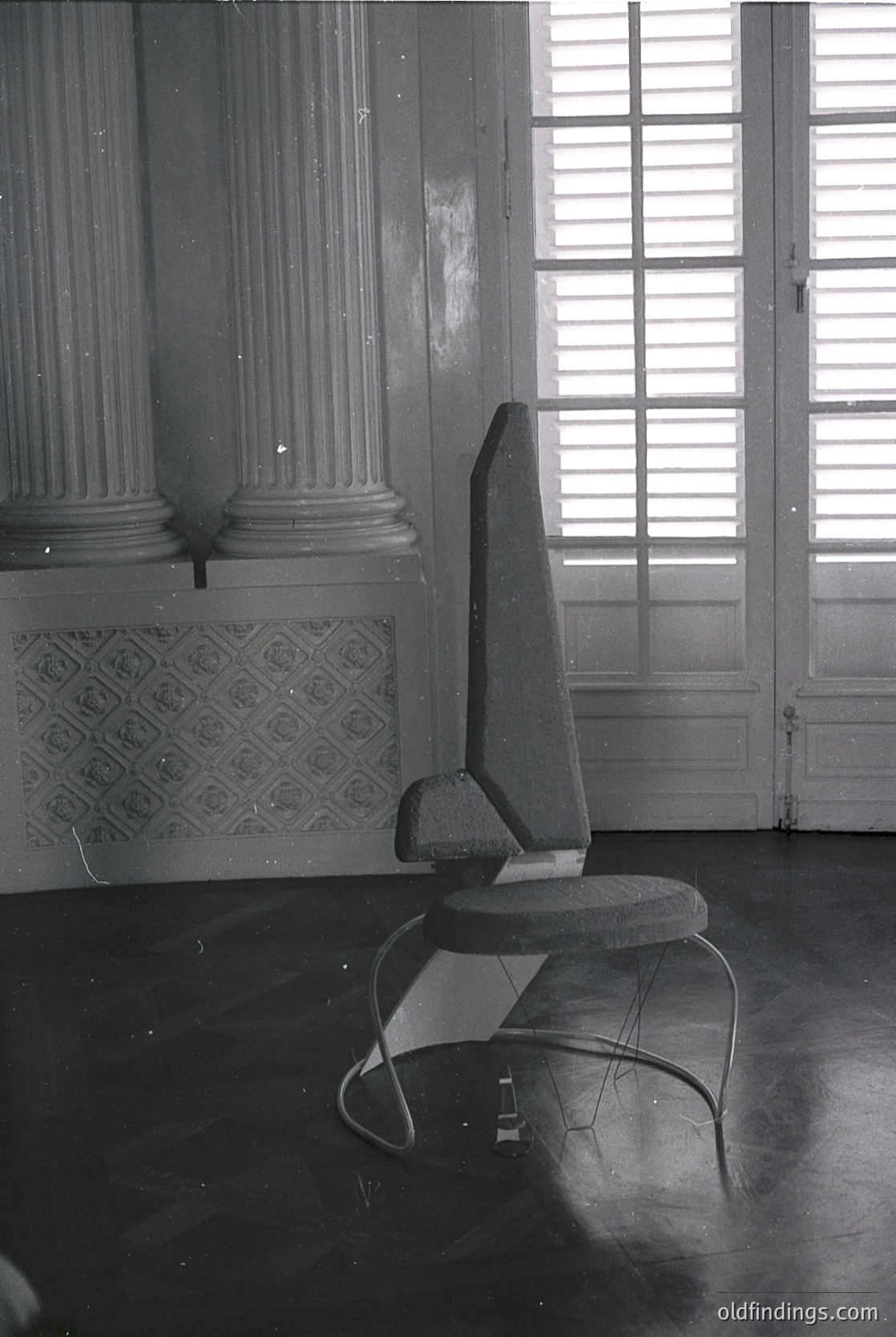 Mid-century modern chair with sculptural metal frame and round seat, positioned in a grand room with classical columns and patterned tile. Natural light floods through tall, arched windows.