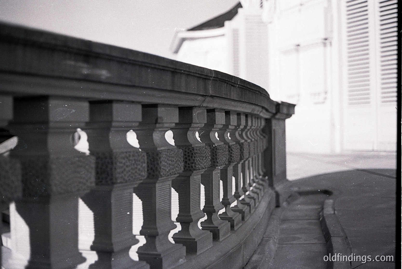 Black-and-white photograph of ornate wrought-iron balustrade with scalloped balusters and curved handrail, reflecting in wet pavement. Architectural detail suggests mid-20th century European influence.